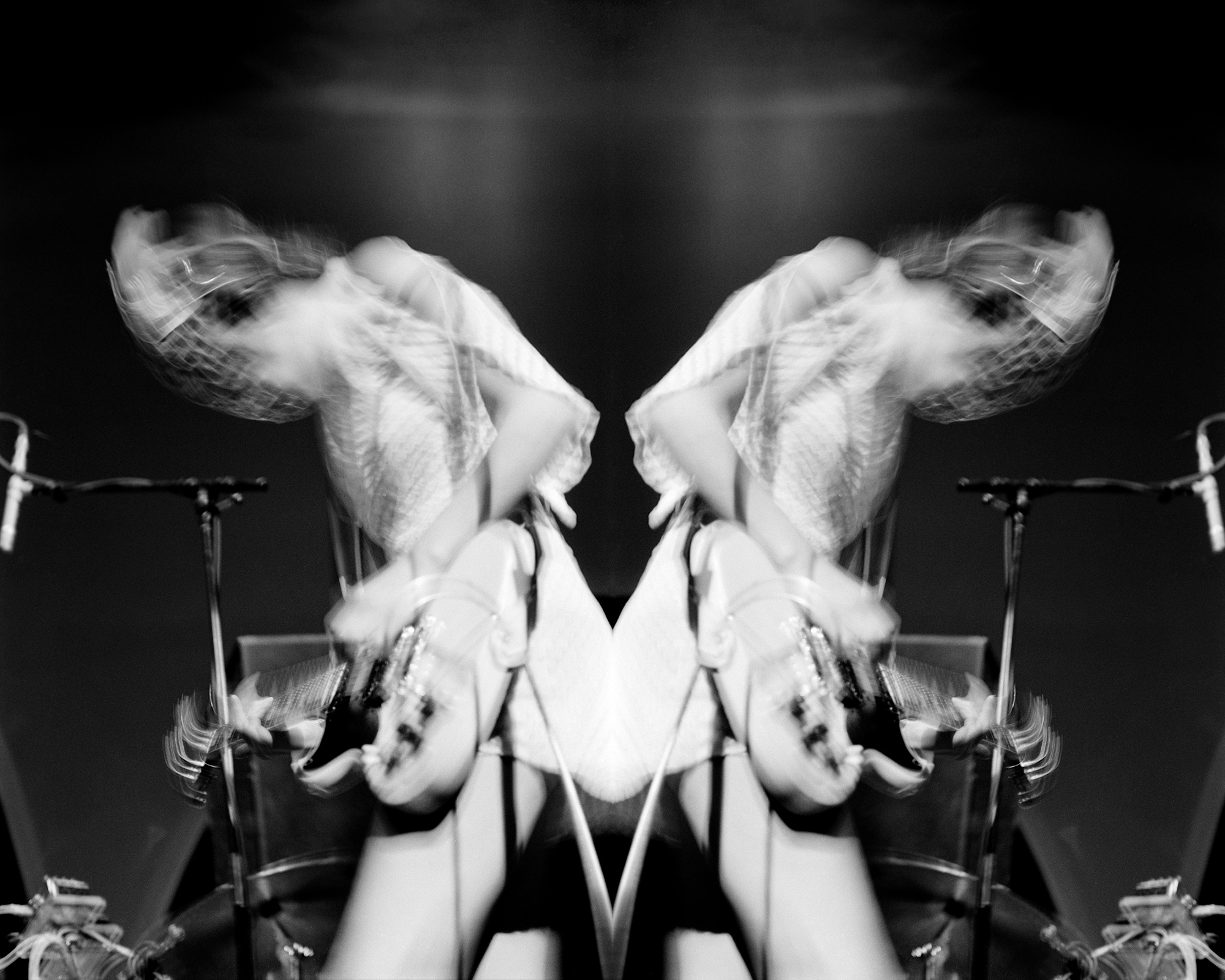 Black and white mirrored image showing blurred motion of hands and arms creating symmetrical white shapes against dark background.
