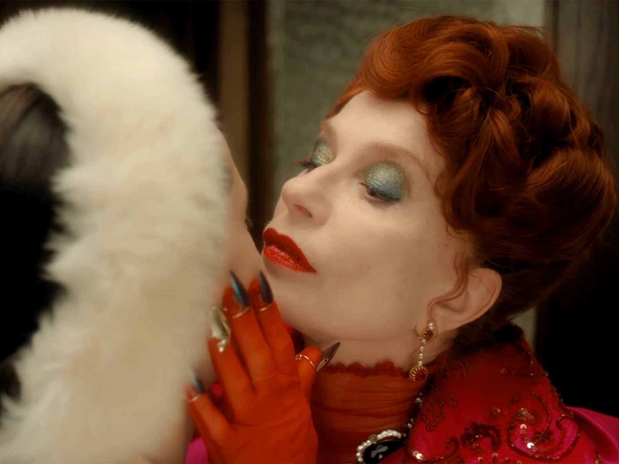 Woman with red curly hair and red lipstick wearing red gloves and clothing, looking up at white feathered object against dark background.