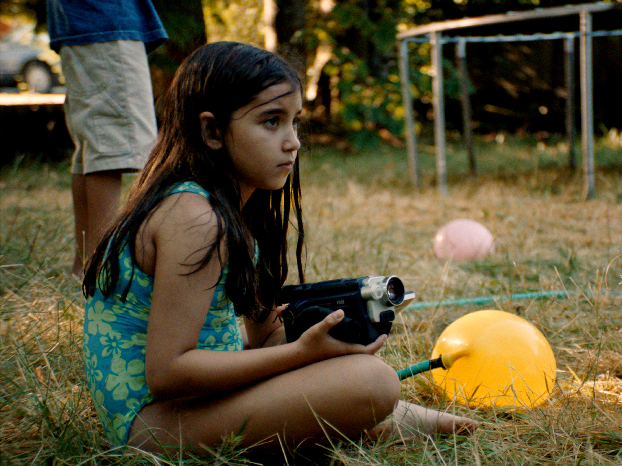Young girl in blue dress sits on grass holding black device, with orange and pink balloons nearby and football goal in background.