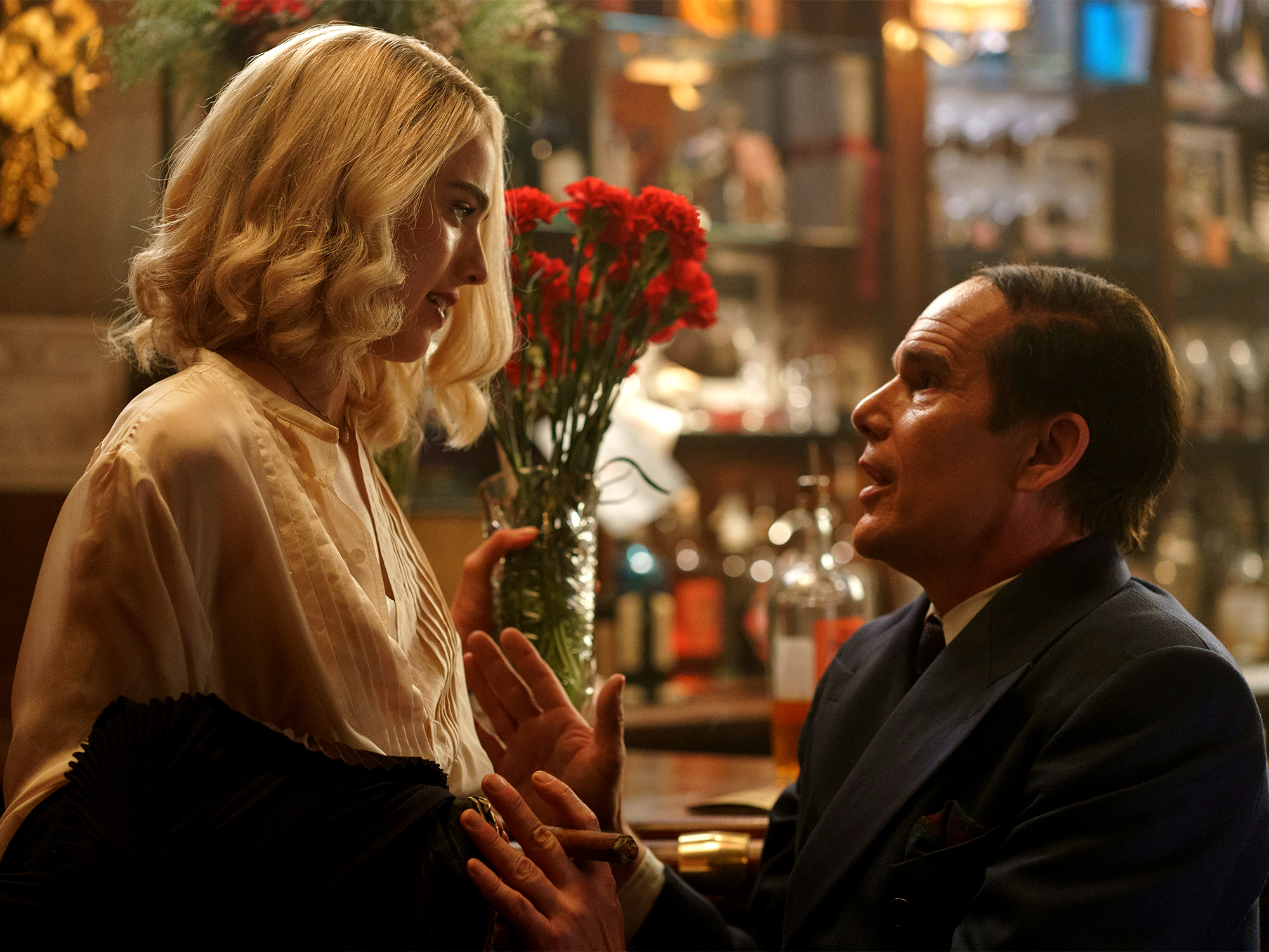 Blonde woman in cream blouse receives red roses from kneeling man in dark suit inside warmly lit restaurant or pub setting.