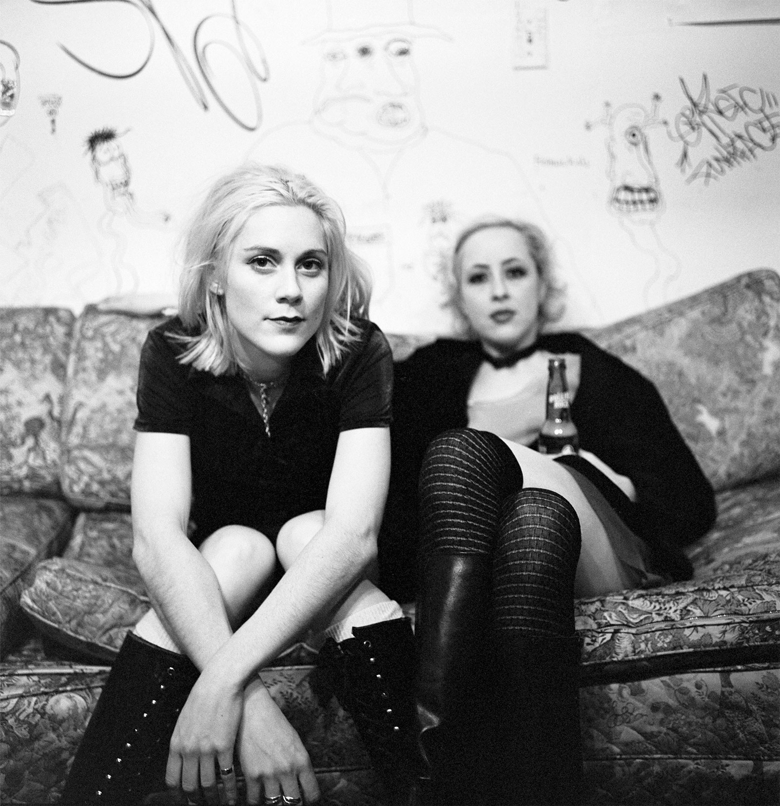 Two women in black clothing sitting on patterned sofa. Black and white image with graffitied wall behind them.
