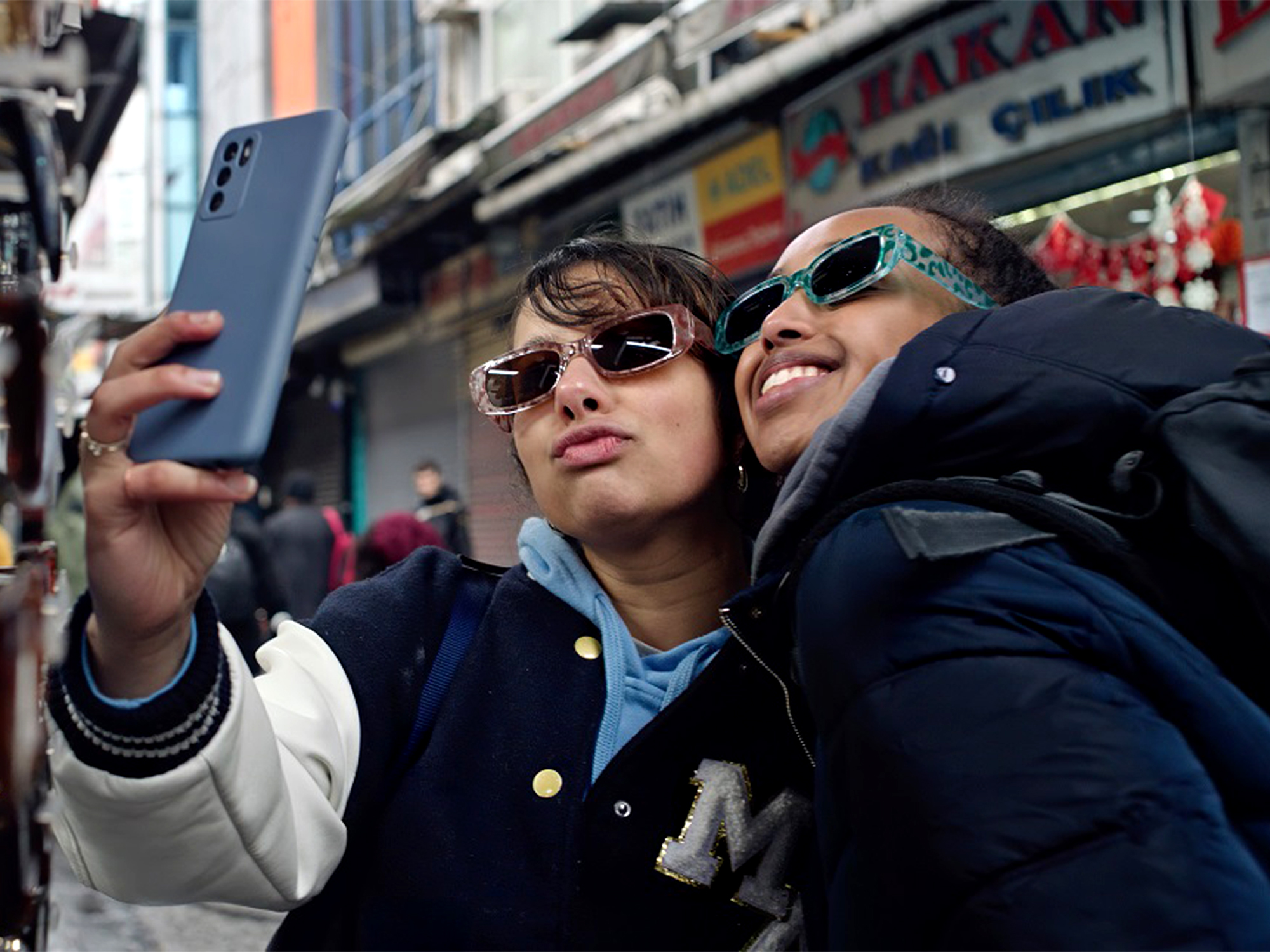 Two young women wearing sunglasses take a selfie on a busy street with colourful shop signs in the background.