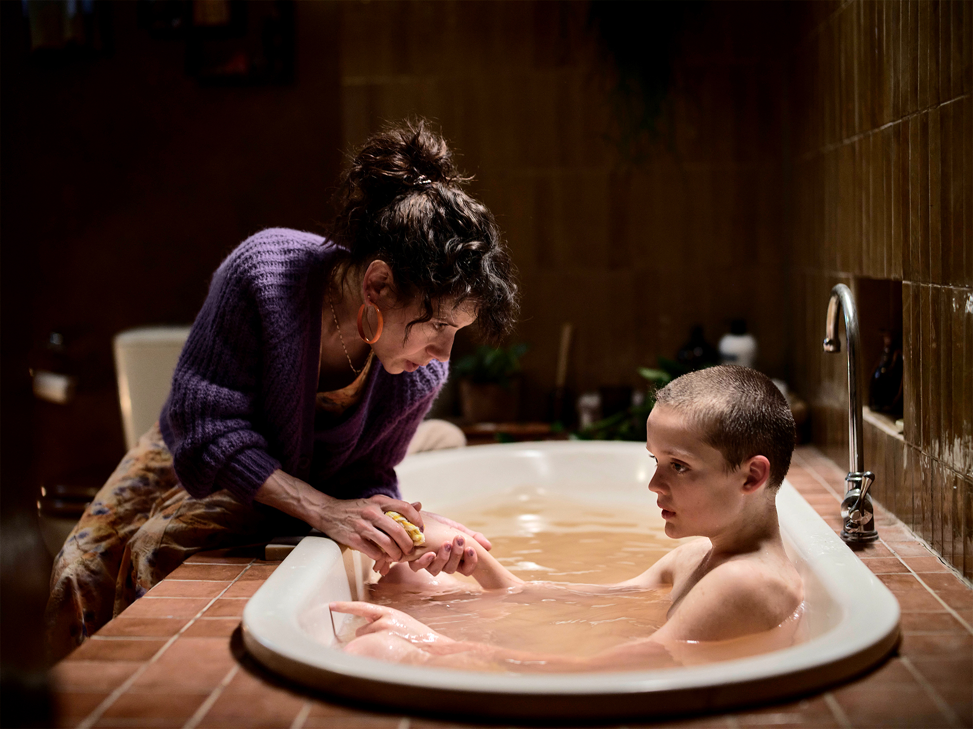 Woman in purple jumper leaning over white bathtub, assisting person with short hair bathing in warm-lit bathroom setting.
