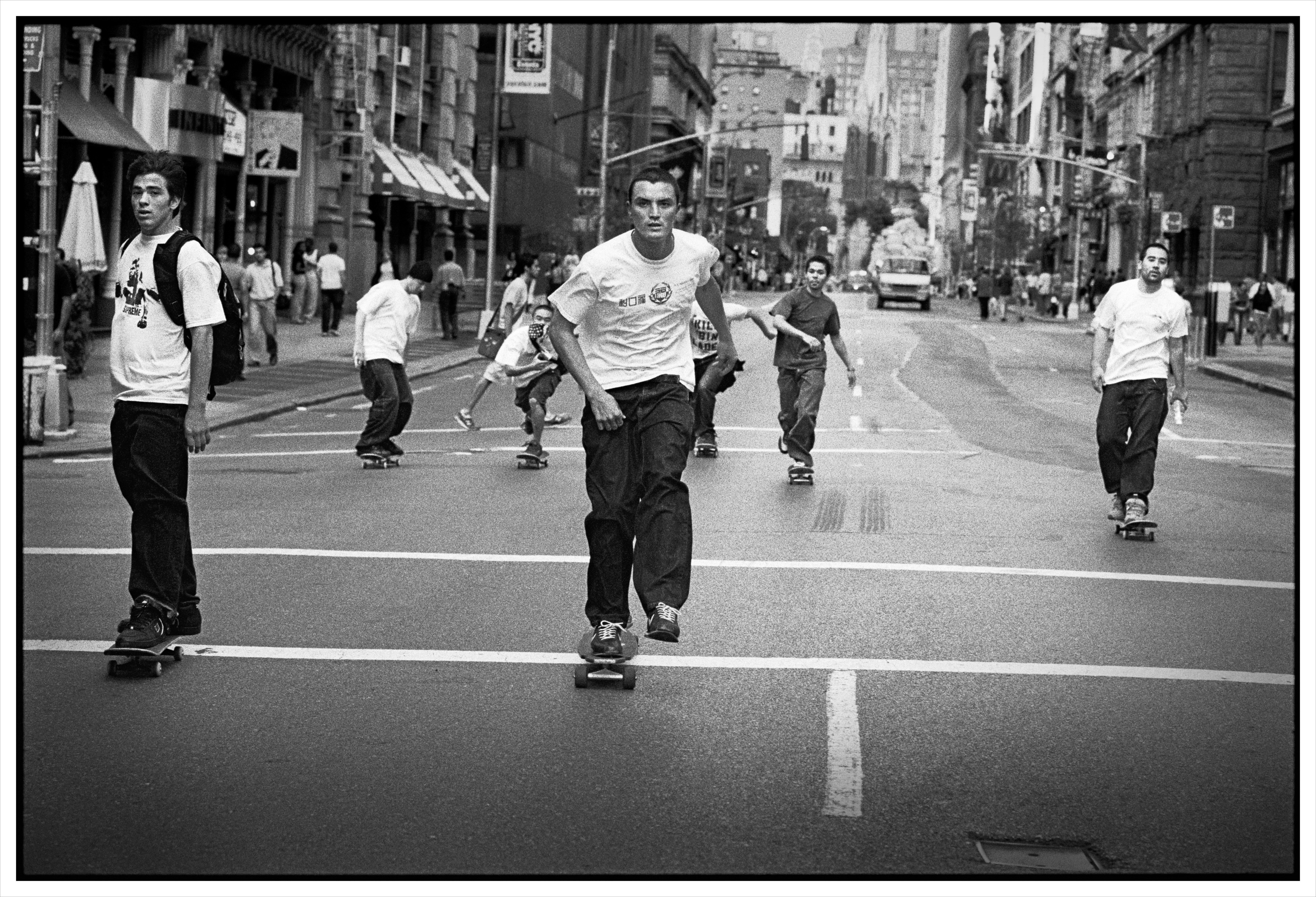 Black and white street scene with several people roller skating down a road between urban buildings and storefronts.