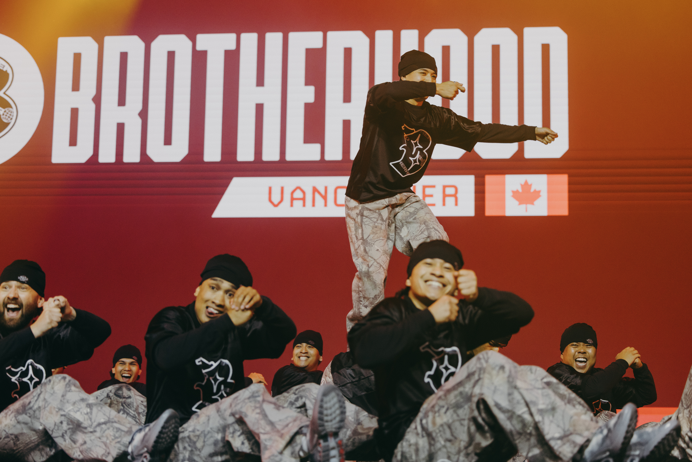 Dance crew performing on stage with red backdrop featuring "BROTHERHOOD" text and Vancouver Canadian flag logo.