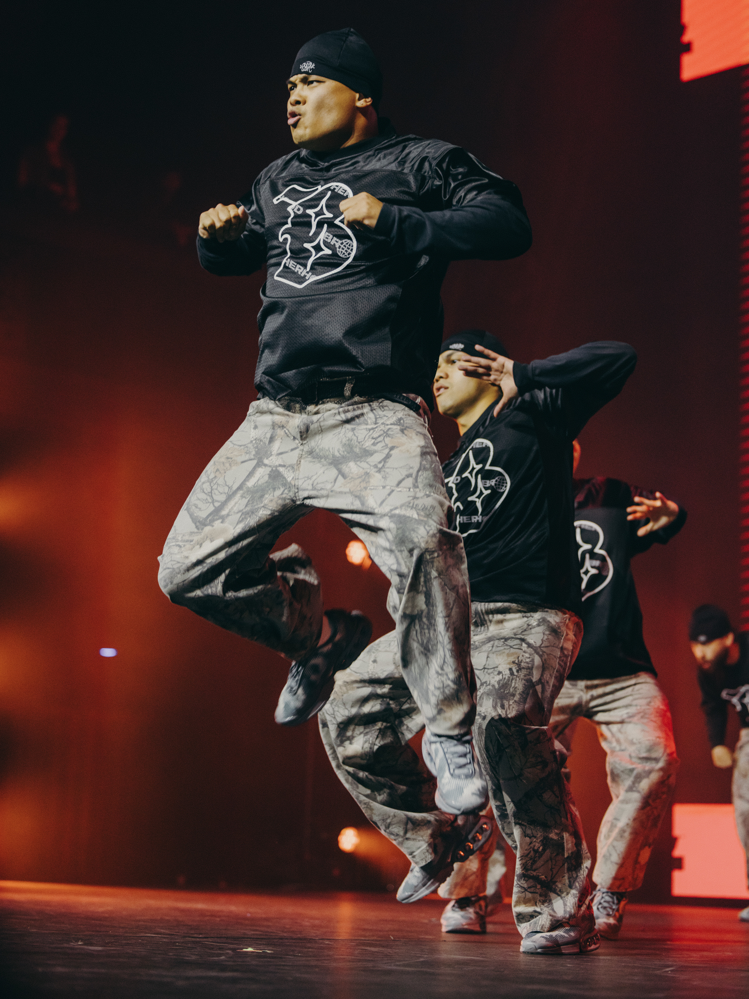 Two dancers in black jumpers and grey camouflage trousers performing on stage with red lighting and smoke effects in background.
