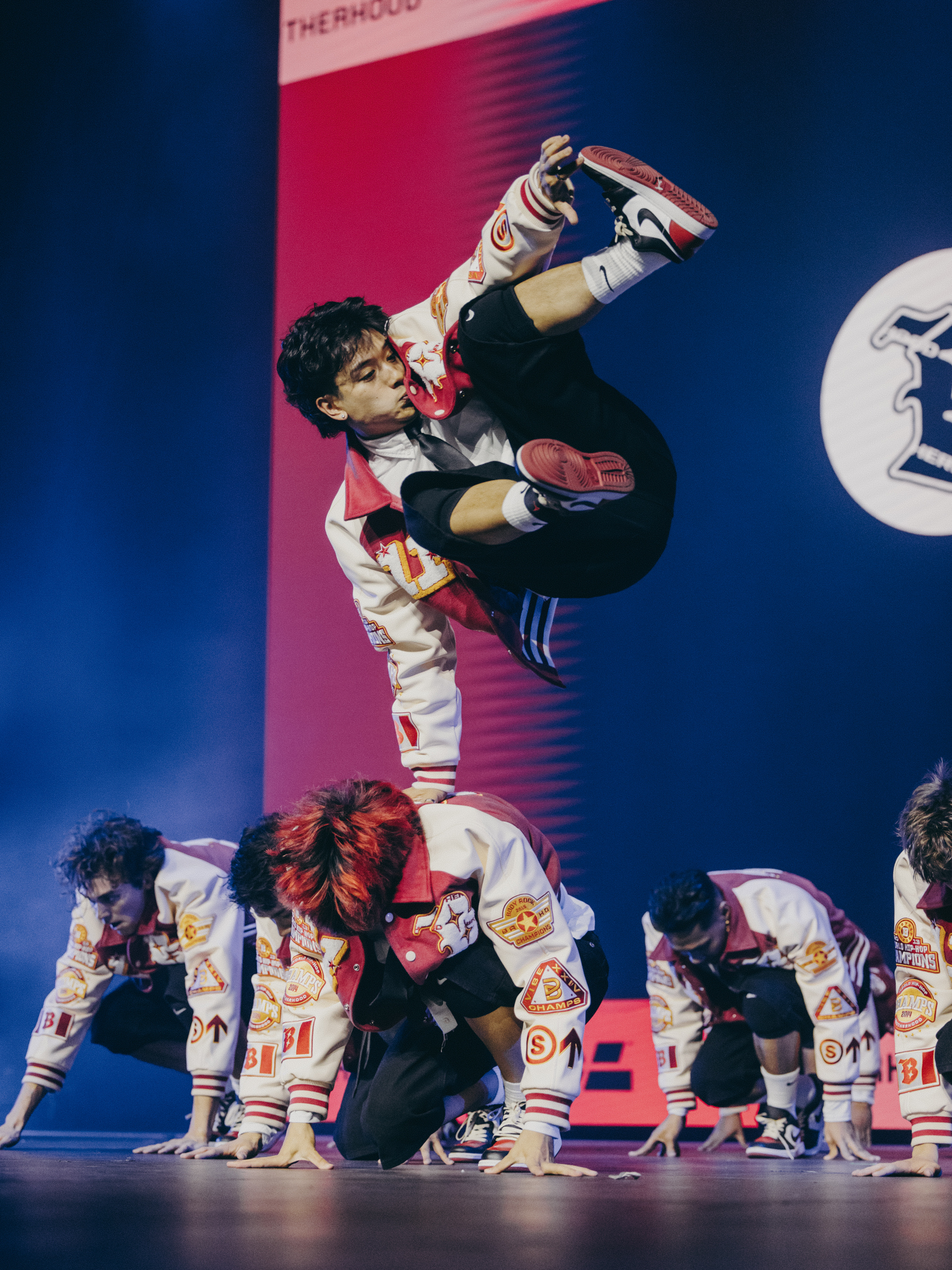Taekwondo athlete performing high kick above group of kneeling competitors on stage with pink and blue backdrop.