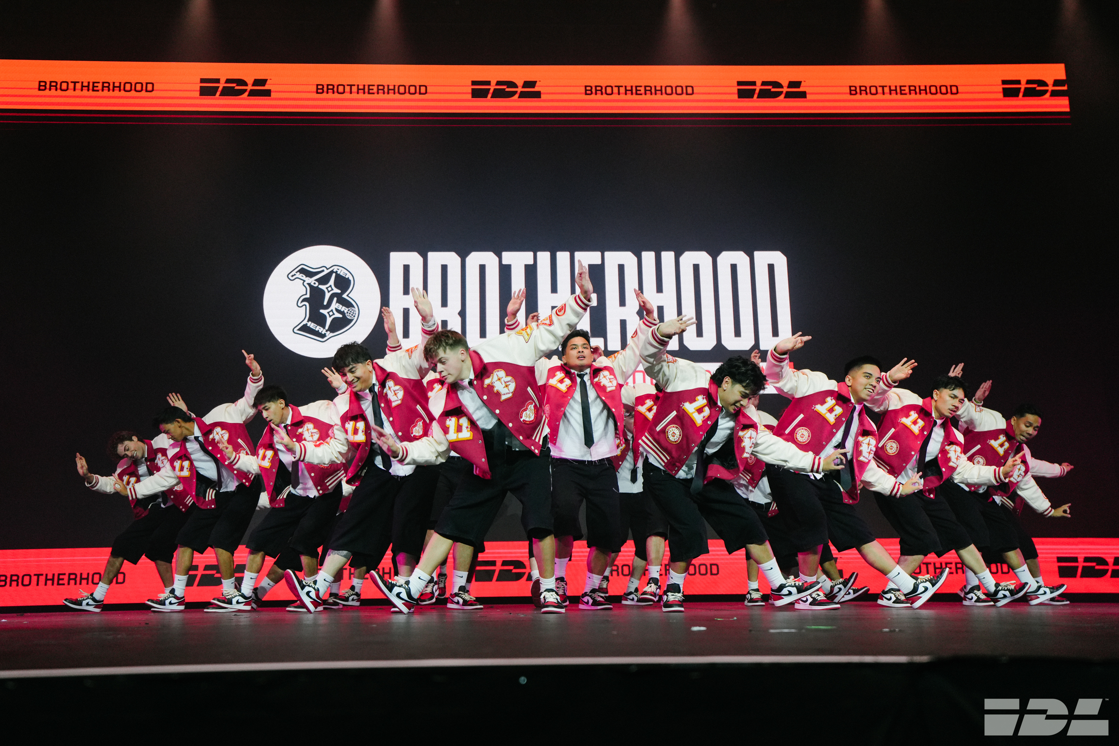 Dance group in red and white uniforms performing on stage with "BROTHERHOOD" logo backdrop in black, white and red colours.