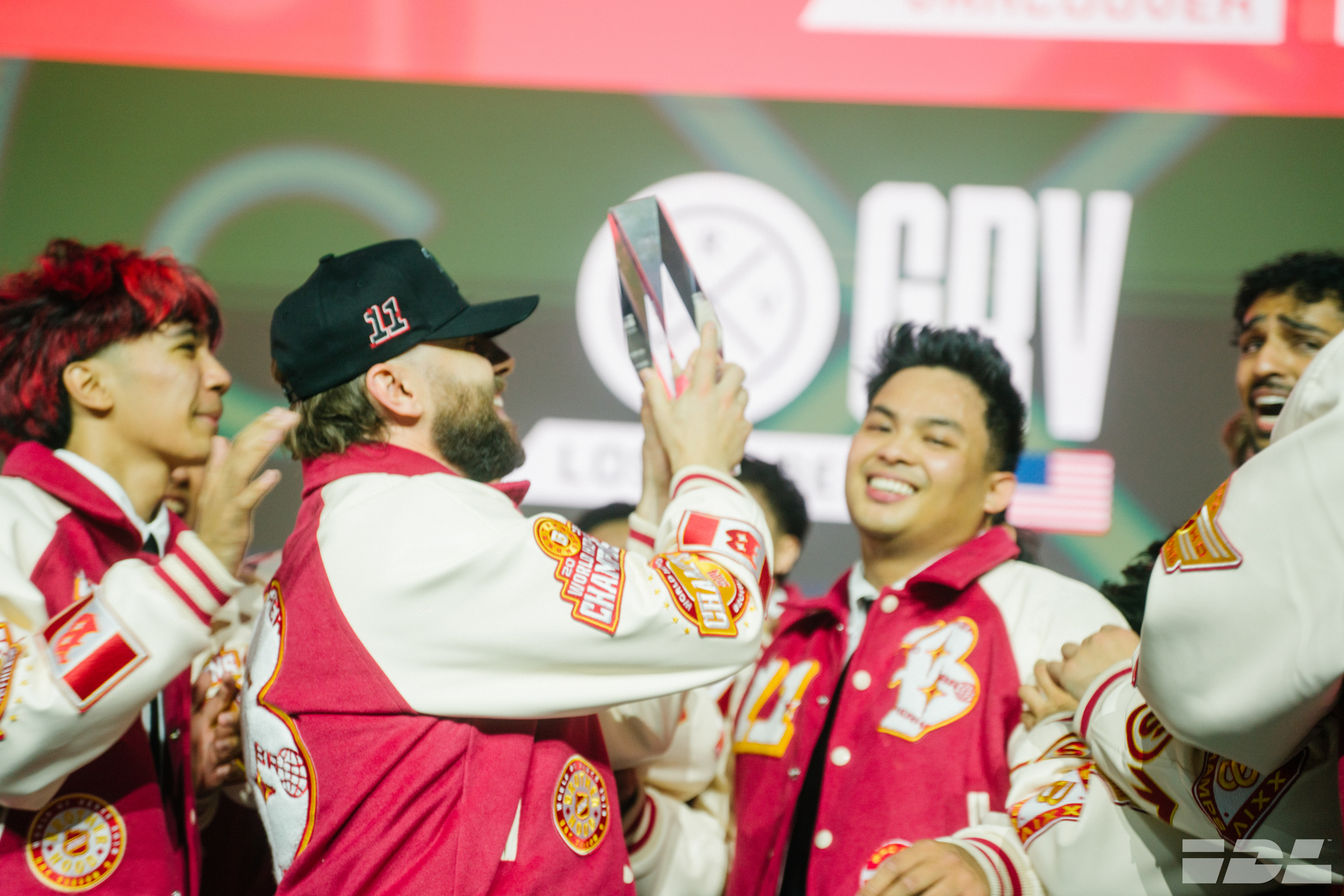 Five young men in red and white sports uniforms celebrating together, with one wearing a black cap, against a green and red background.