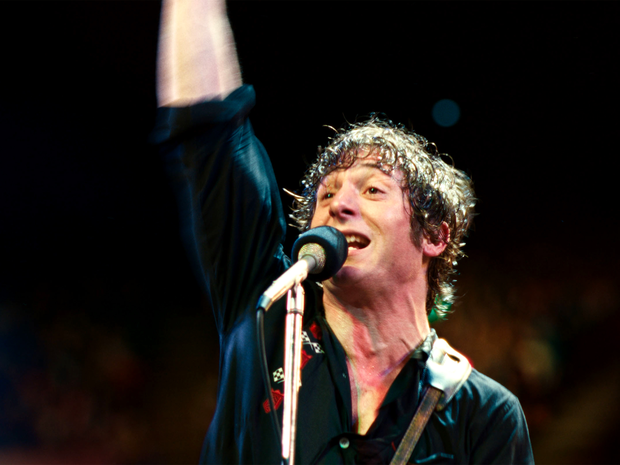 Male performer singing into microphone with raised arm, wearing dark jacket, curly hair, against black background.