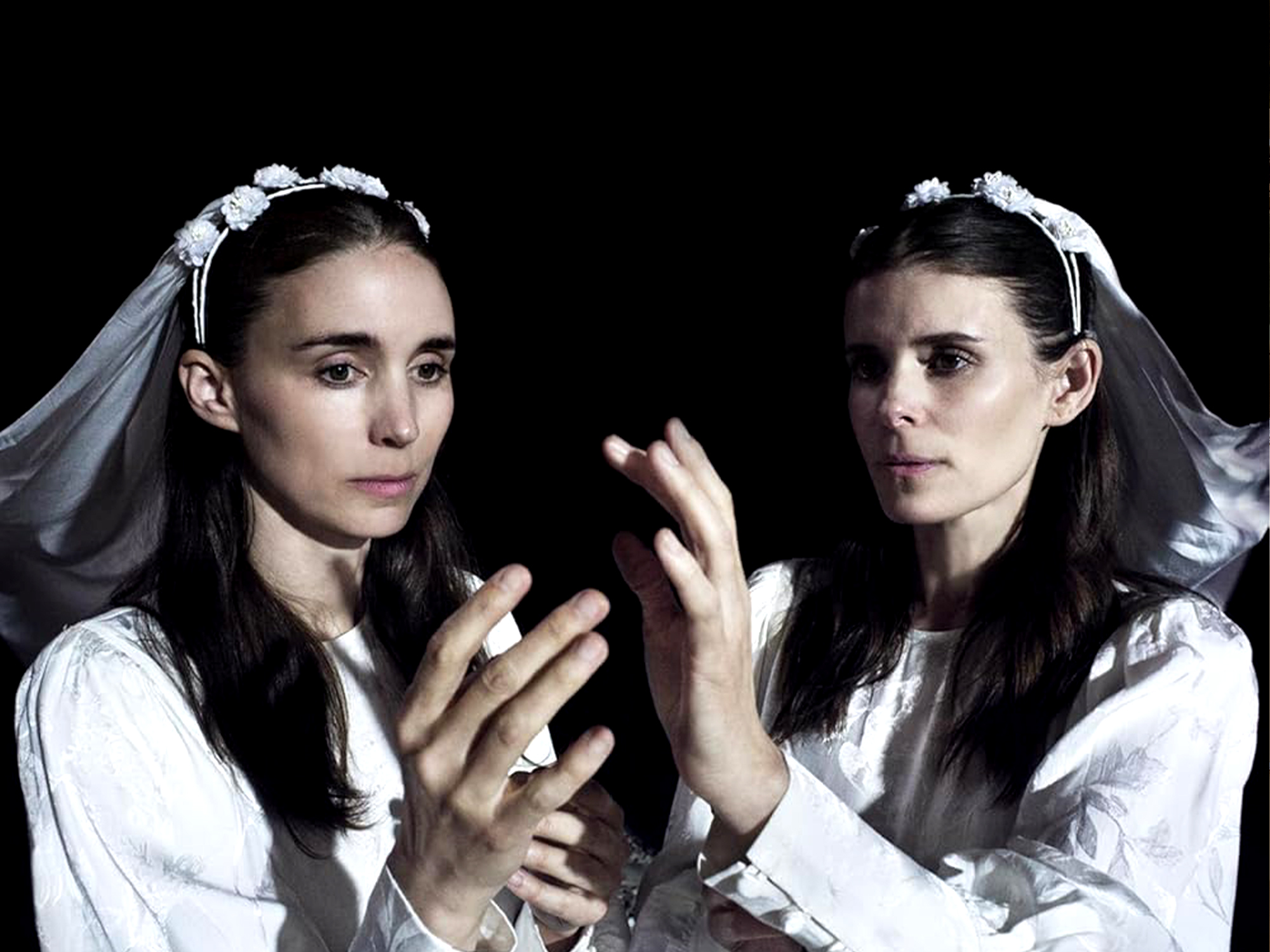 Two women in white wedding dresses and veils with hands pressed together against black background, dark hair, dramatic lighting.