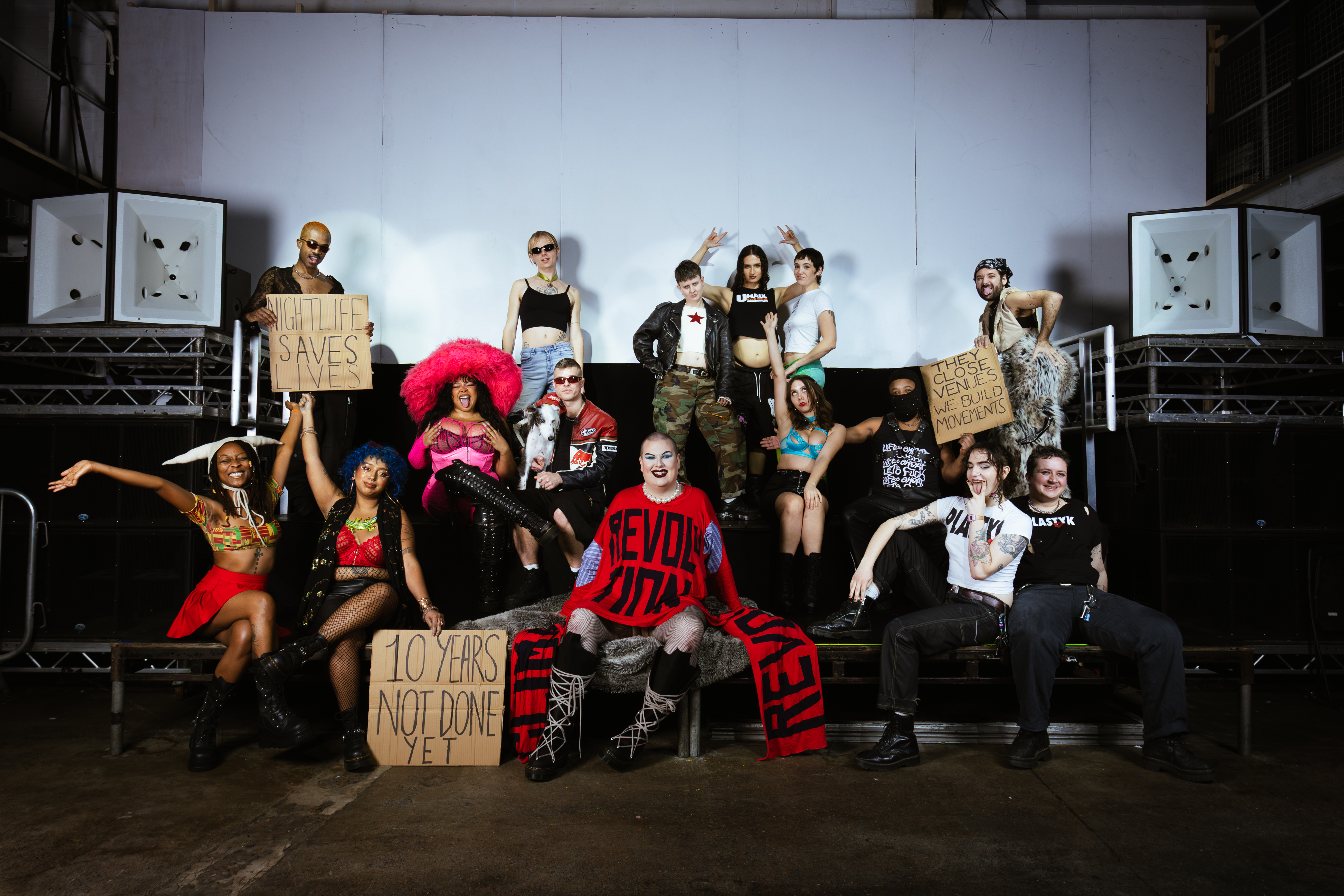 Group of young people posing on black metal stairs holding cardboard protest signs, wearing casual clothing against white backdrop.