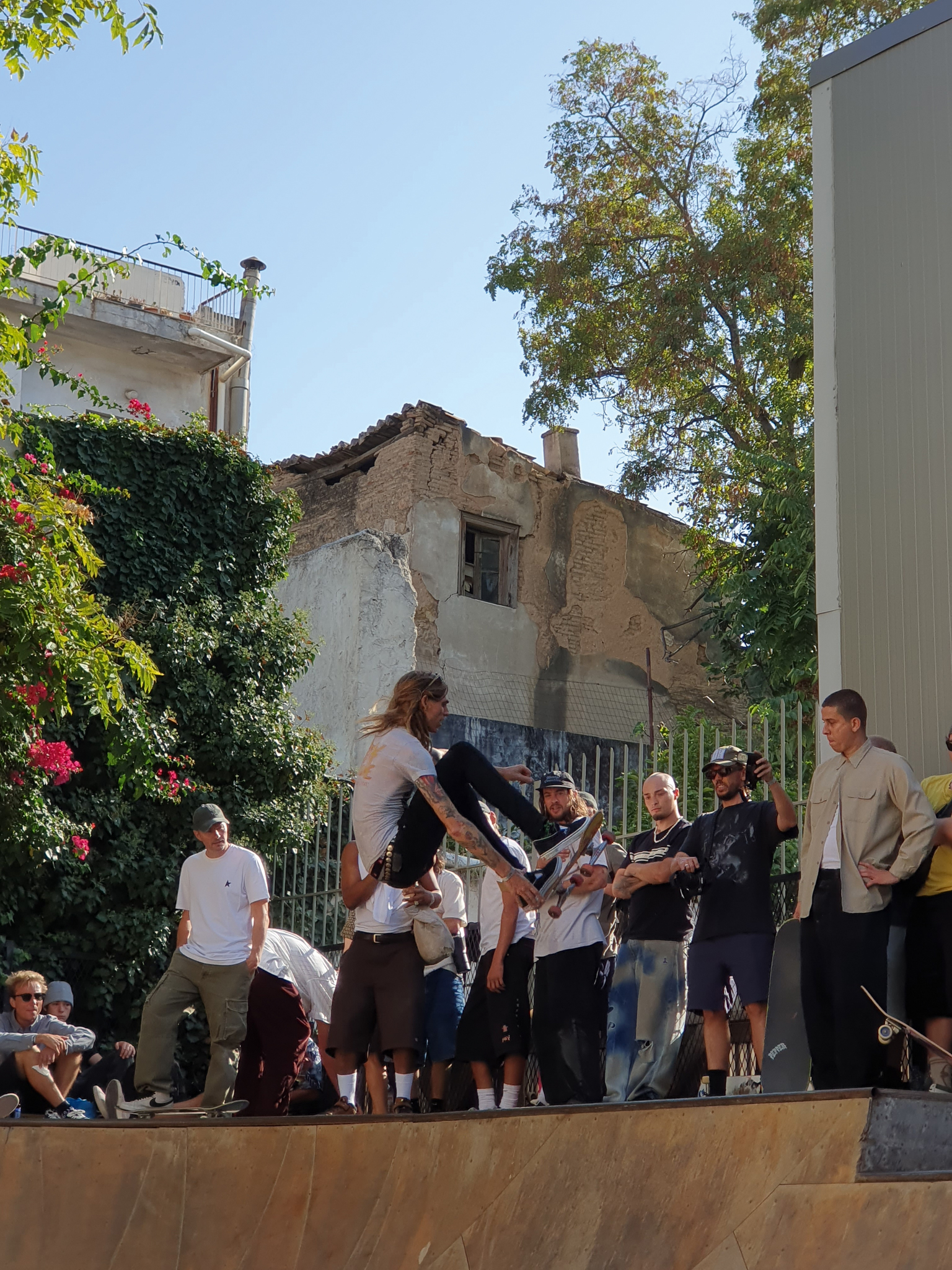 Skateboarder performing trick whilst crowd watches in urban courtyard with damaged building and green foliage in background.