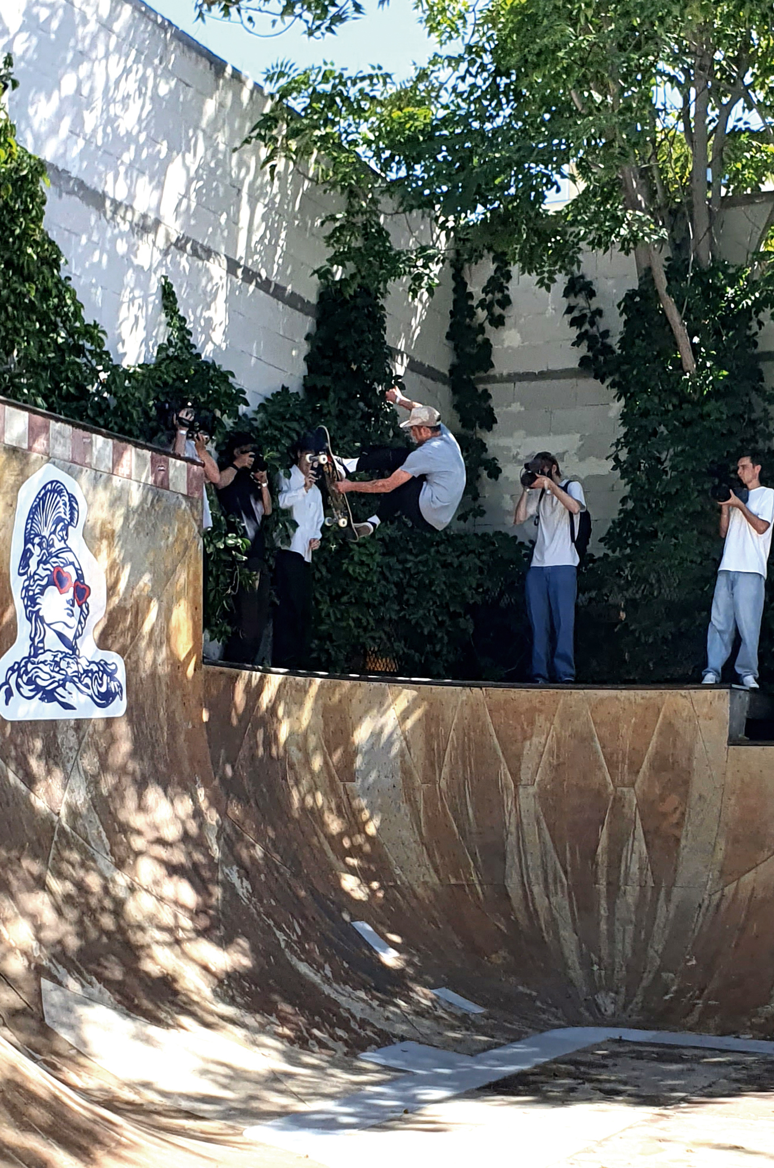 Skateboarder in concrete bowl with spectators and cameraman above, leafy trees casting shadows, blue portrait artwork on wall.