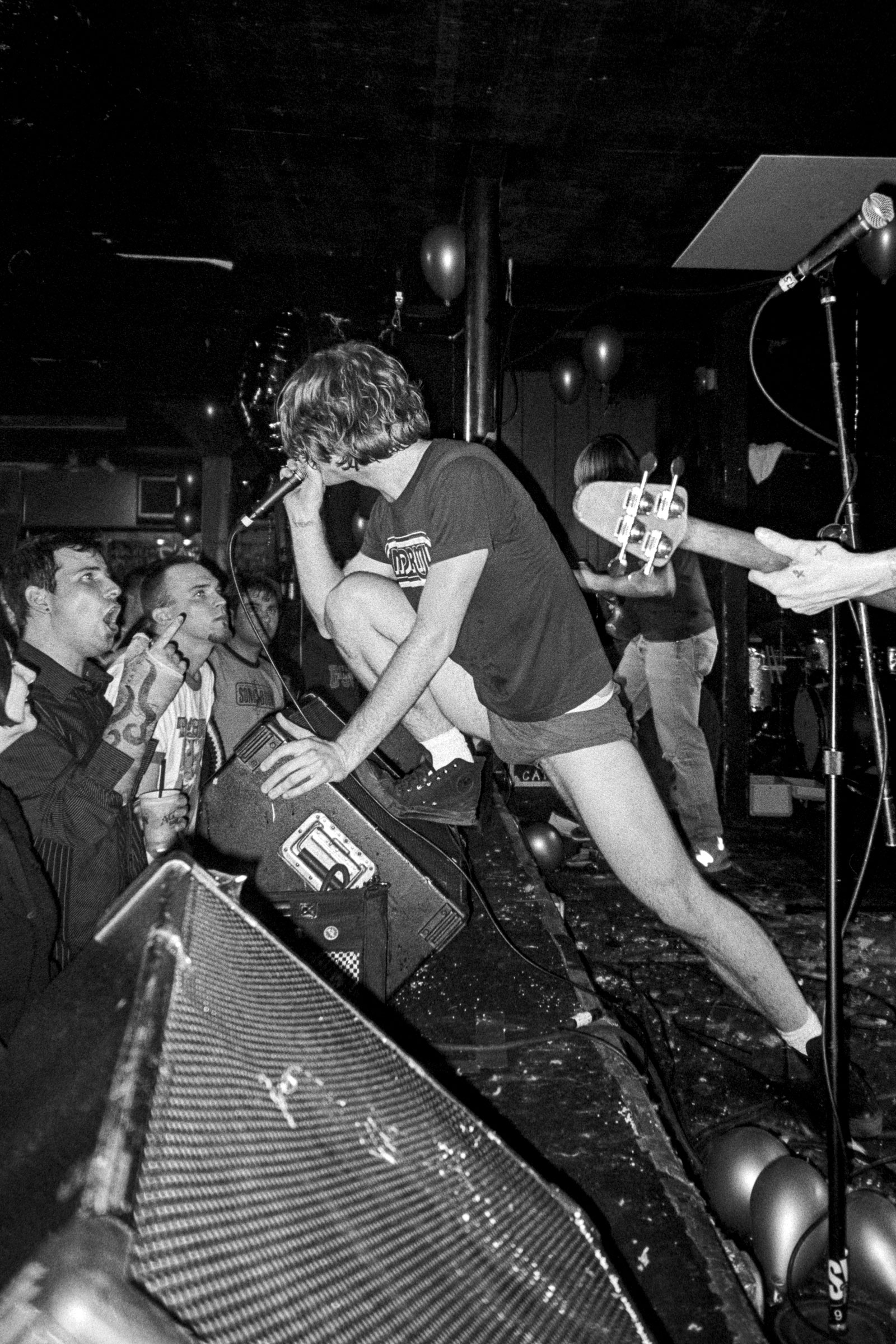 Black and white image of punk rock performance with vocalist leaning into crowd, amplifier visible, audience watching from left side.
