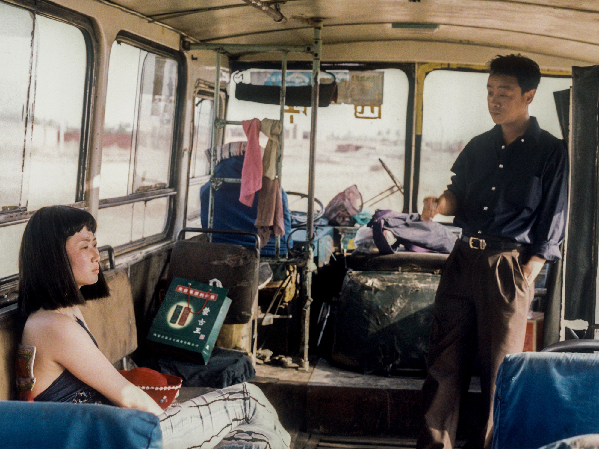 Cluttered interior of a van, with a woman sitting on a couch and a man standing. Varied colours and shapes, with items scattered throughout the space.
