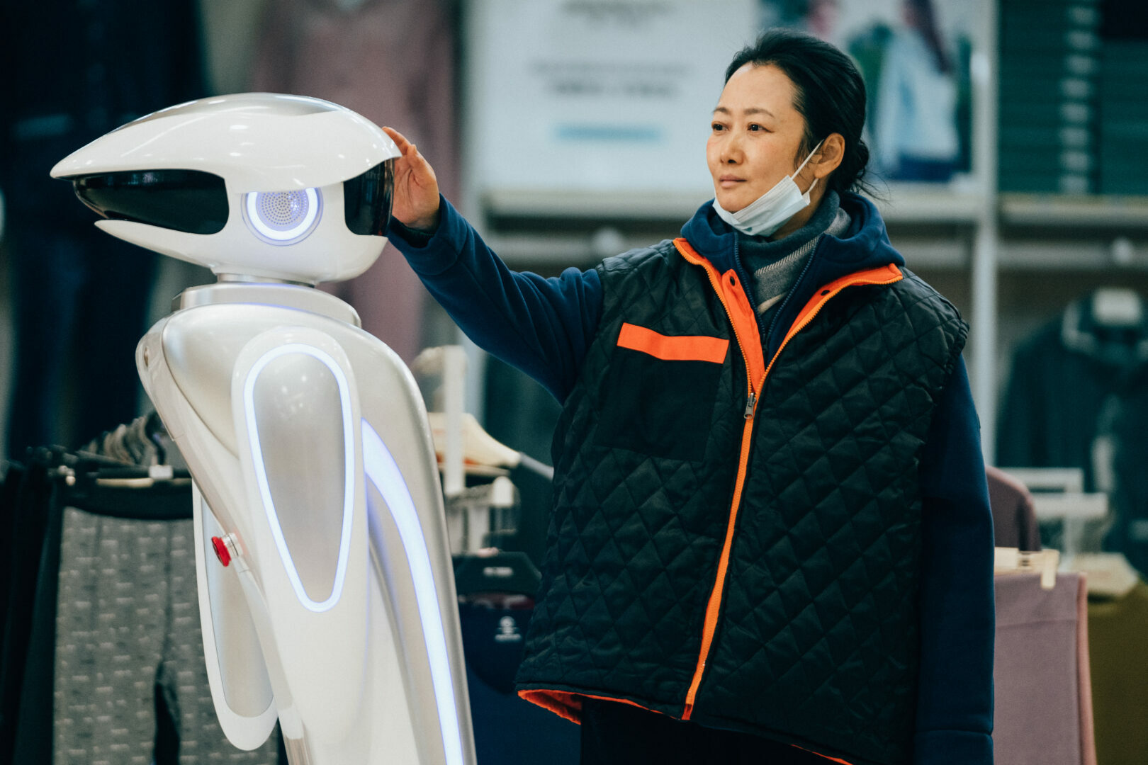A woman in a black and orange coat interacting with a white robot in an indoor setting.