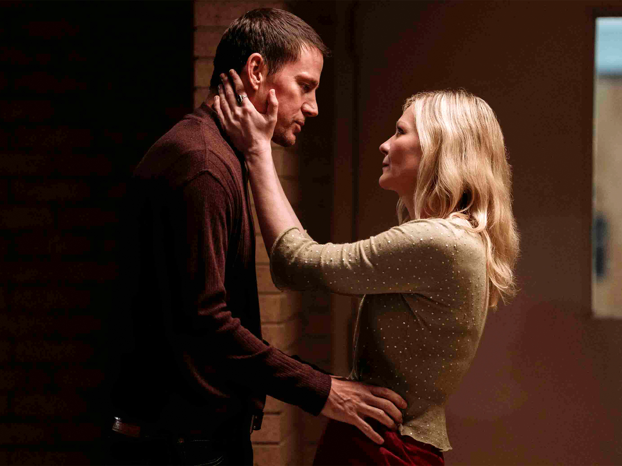 Blonde woman in beige jumper touching face of man in brown shirt, both looking at each other intimately against brick wall.