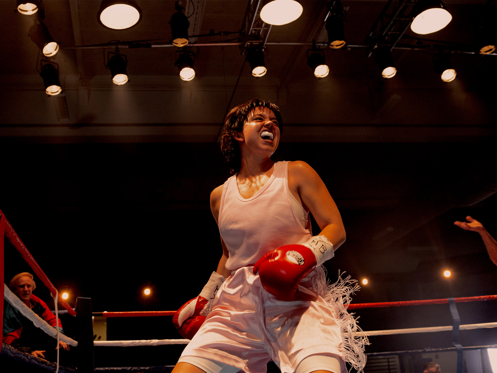 Female boxer in white vest and shorts with red gloves celebrates in boxing ring under bright overhead lights.