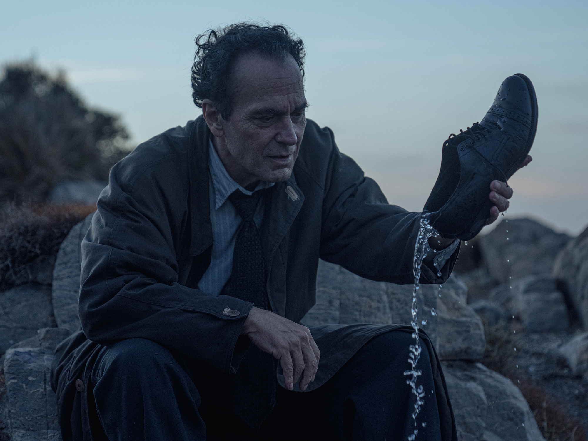 A man in a black suit sits on a rocky cliff, holding a shoe that appears to be dripping water.