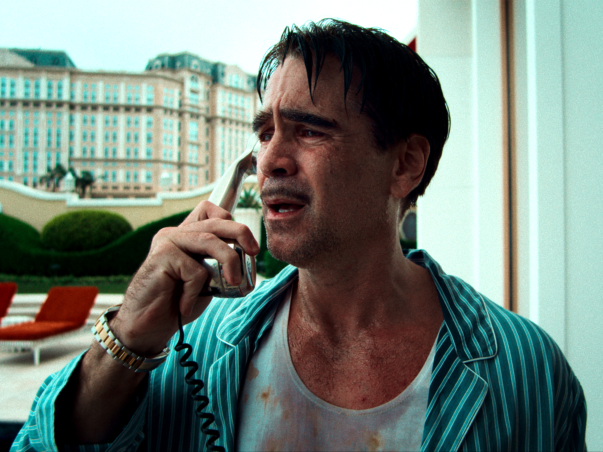 Man in striped shirt speaking on mobile phone, looking distressed with bloodstains on white vest, ornate building visible through window.