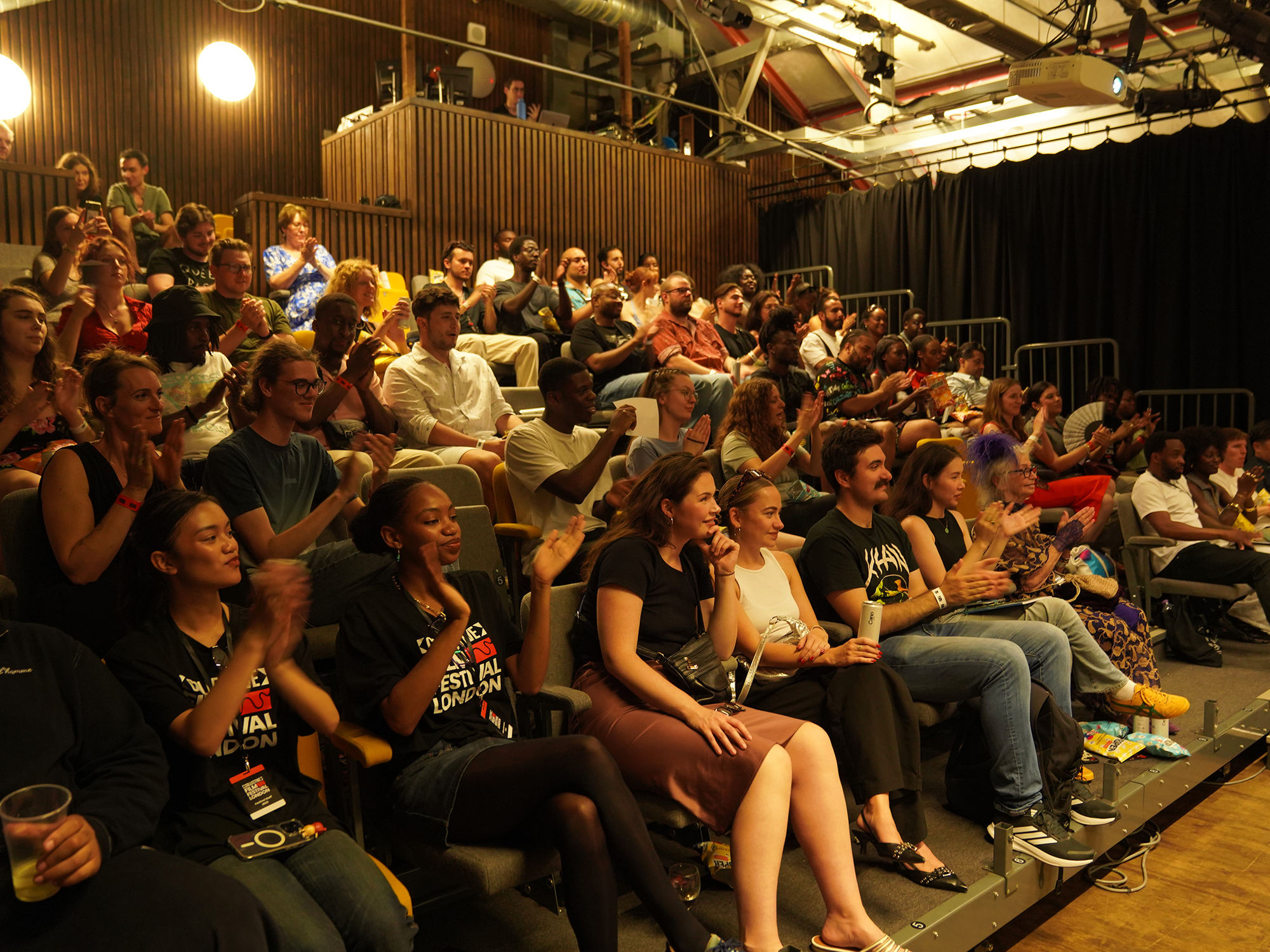 Audience seated in tiered rows of a small theatre or performance space with exposed ceiling beams and warm lighting.