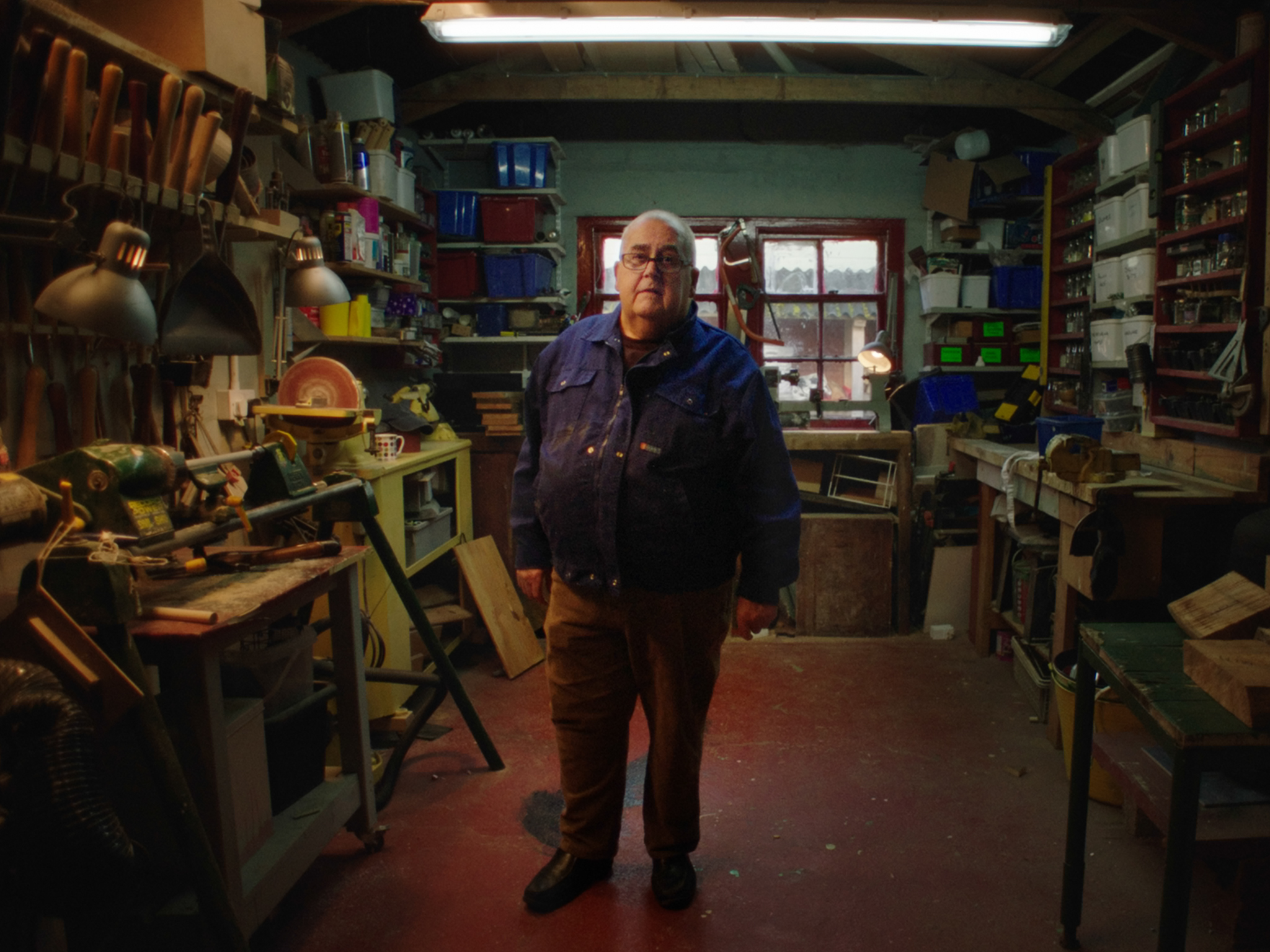 Elderly man in blue jumper standing in cluttered workshop with tools, shelving, and fluorescent lighting overhead.