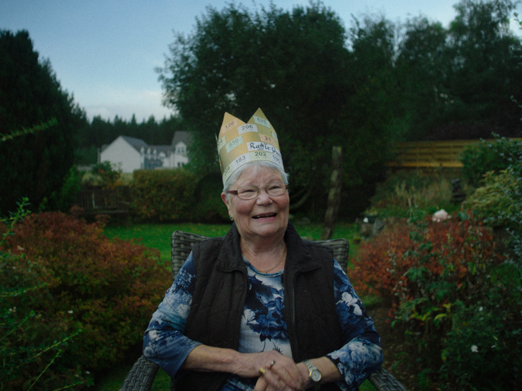Elderly woman wearing paper crown sits in garden chair, smiling. Green trees and houses visible in background, colourful autumn foliage.
