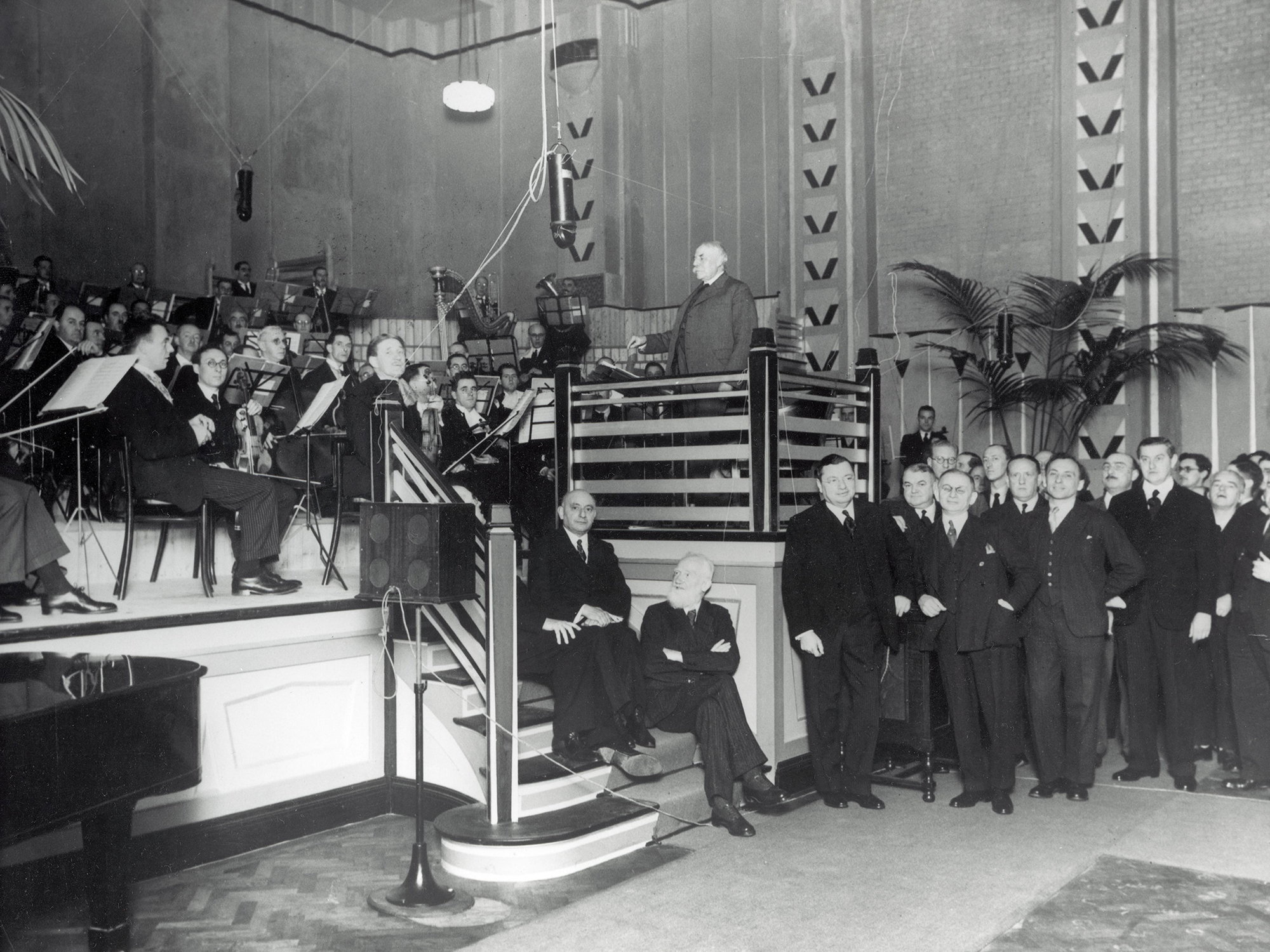 A black and white photograph showing a large group of men in formal attire on stage at Abbey Road's Studio One.