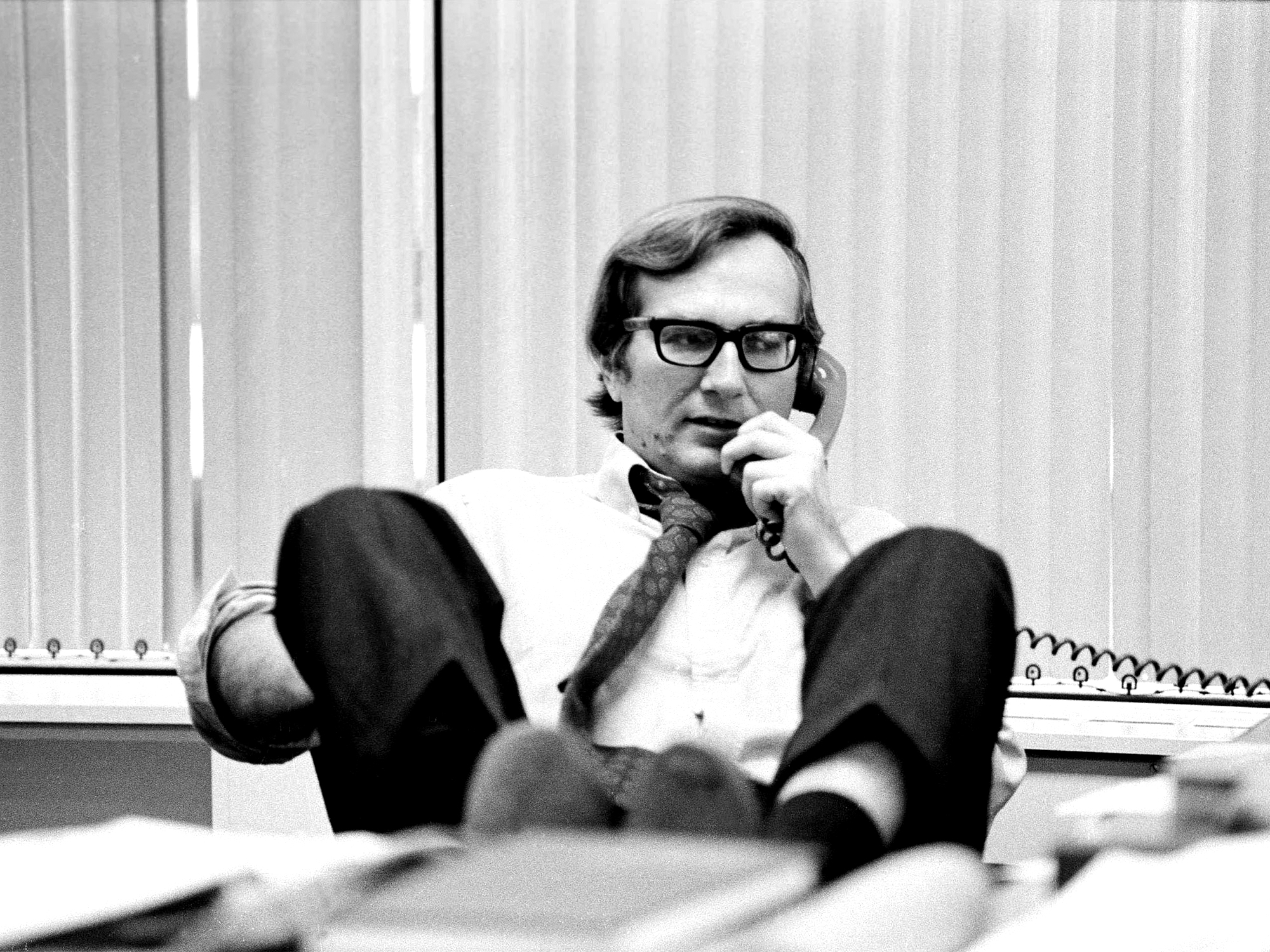 Man in glasses and suit jacket sits at desk with feet up, hand to chin, telephone and papers visible. Black and white image.
