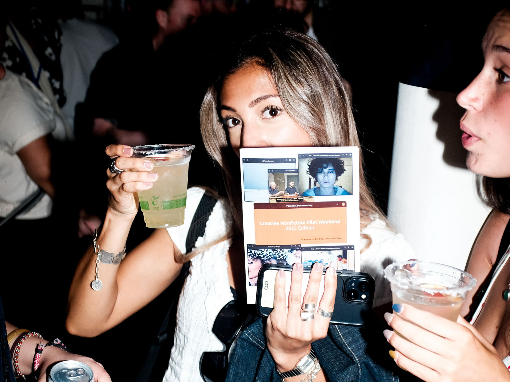Woman with blonde hair holding cocktail and tablet device showing video call, with another woman visible on right holding drink.