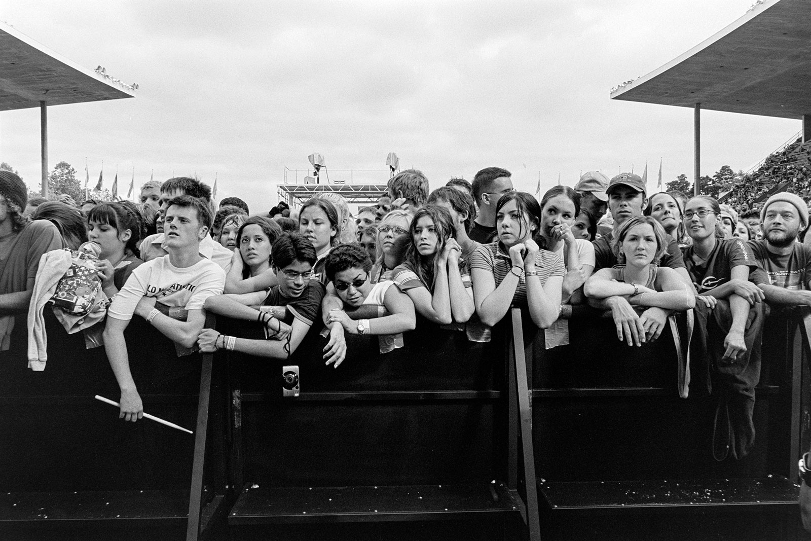 Black and white image of crowd gathered behind metal barriers at outdoor event, with covered structures visible in background.