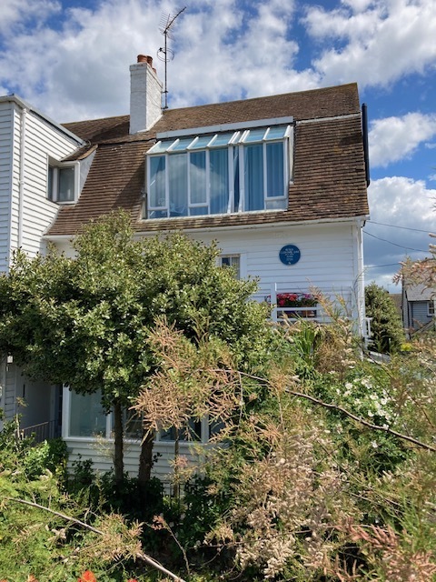 A two-storey home with a brown tiled roof, large windows, white exterior walls and an overgrown garden.