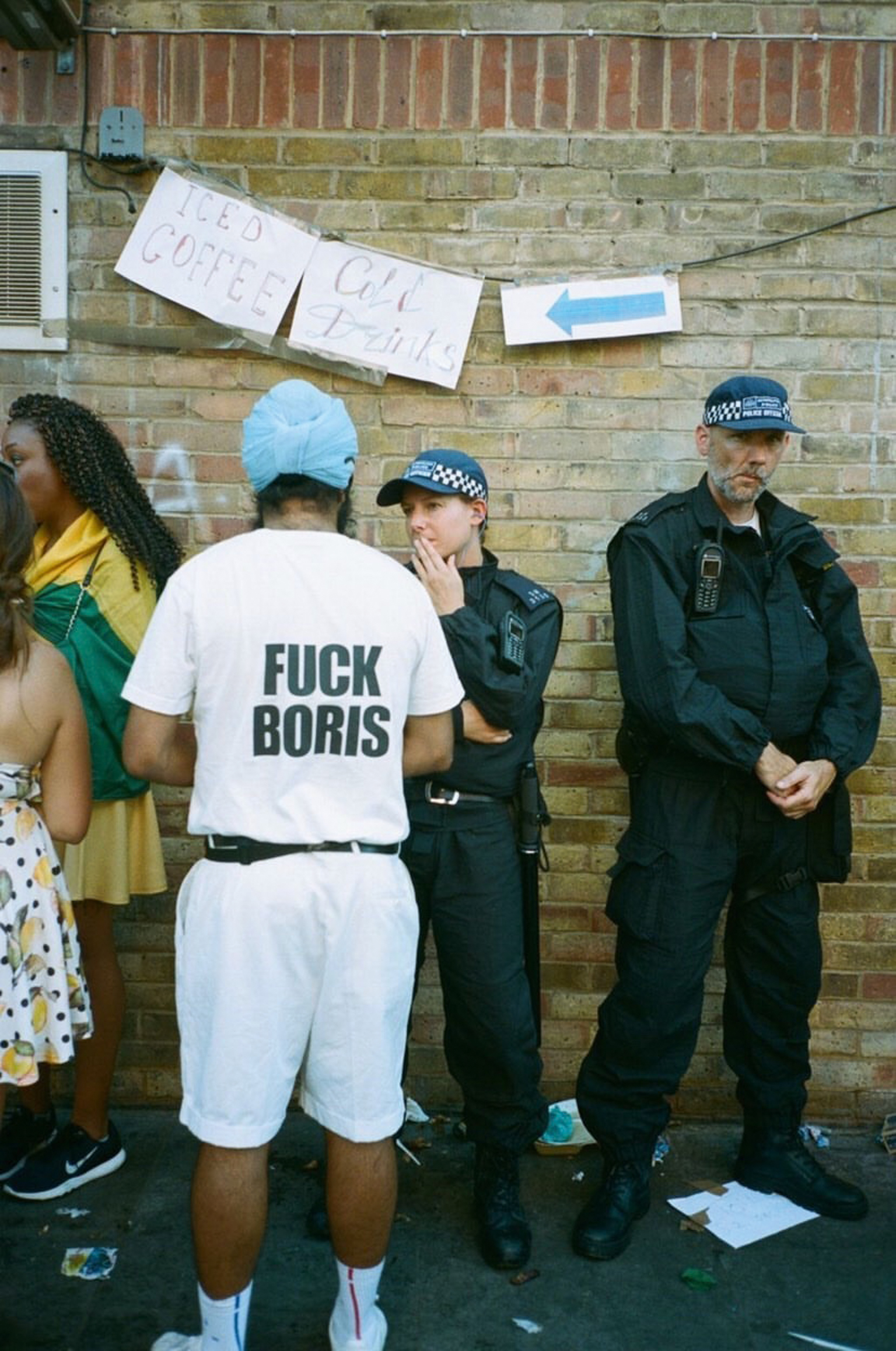 Man in white t-shirt with profanity about Boris Johnson stands near two police officers against brick wall with protest signs hanging above.