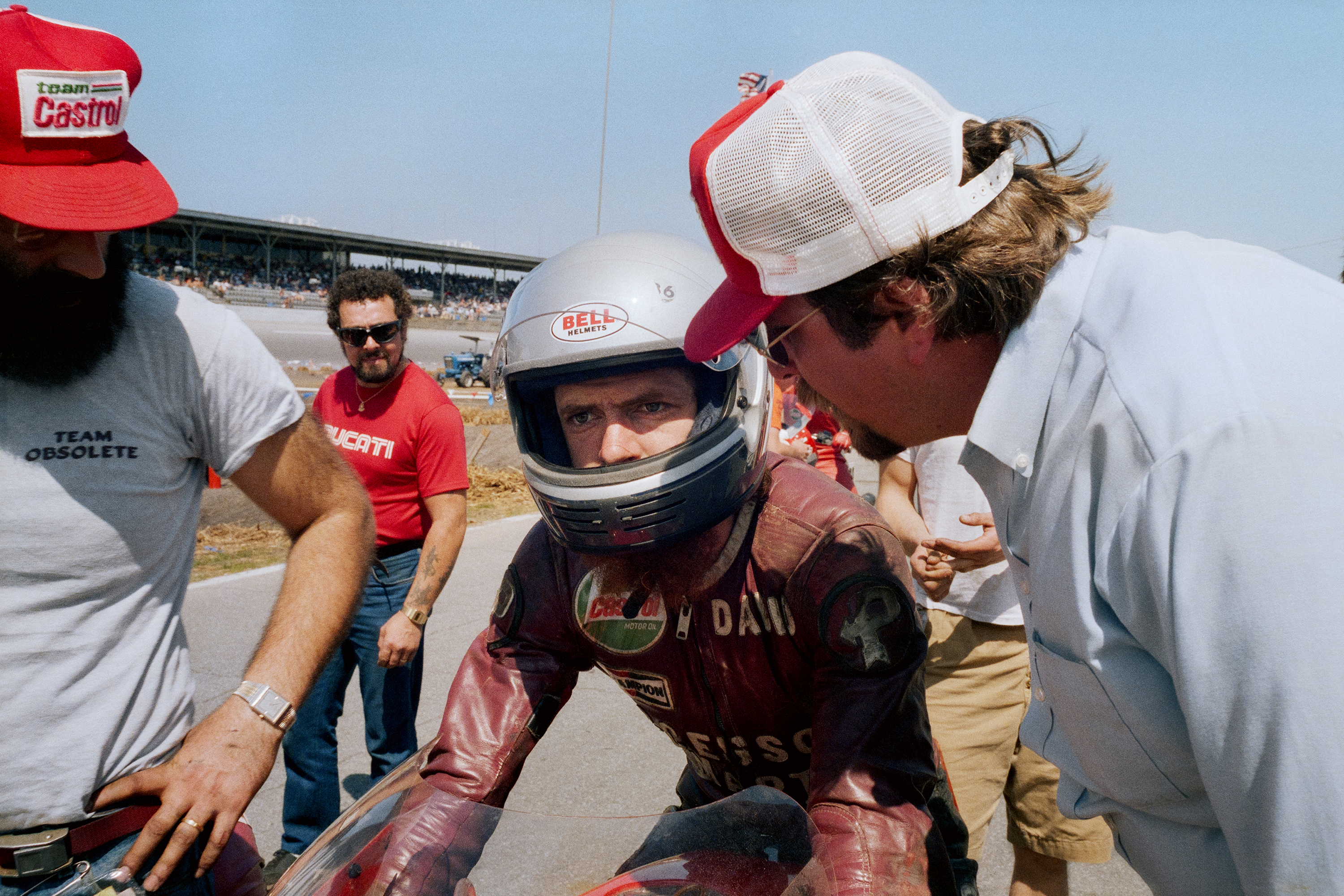 Racing driver in red helmet and suit surrounded by crew members in white caps at sunny racetrack with grandstand visible.