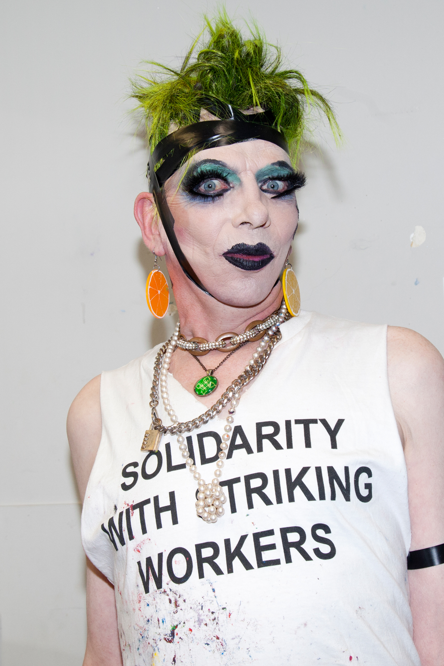 Person in drag makeup with green hair, dramatic eye makeup, wearing white vest reading "SOLIDARITY WITH STRIKING WORKERS" and chains.