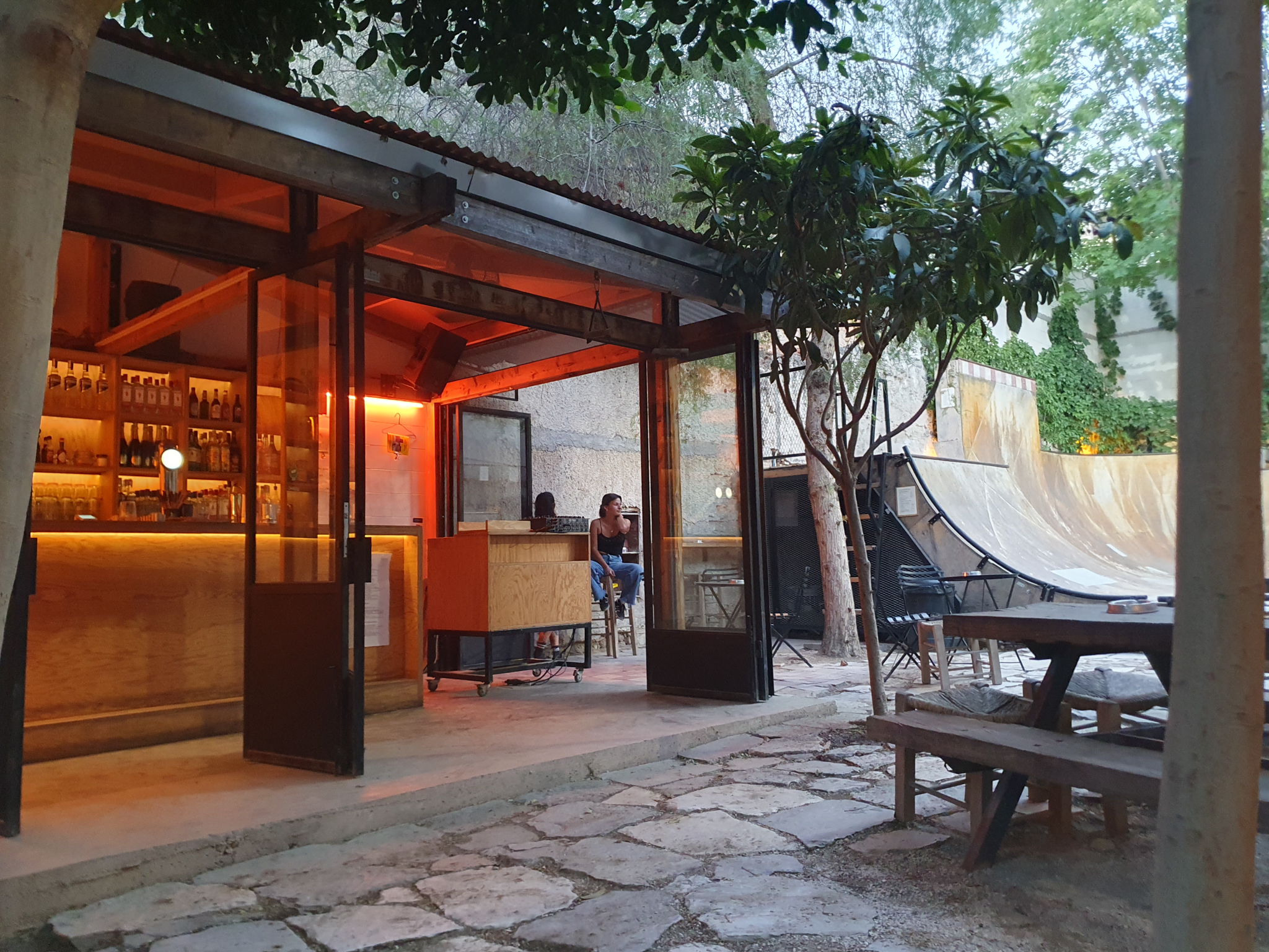 Modern glass-fronted building with warm orange interior lighting, surrounded by trees and paved courtyard with wooden benches.