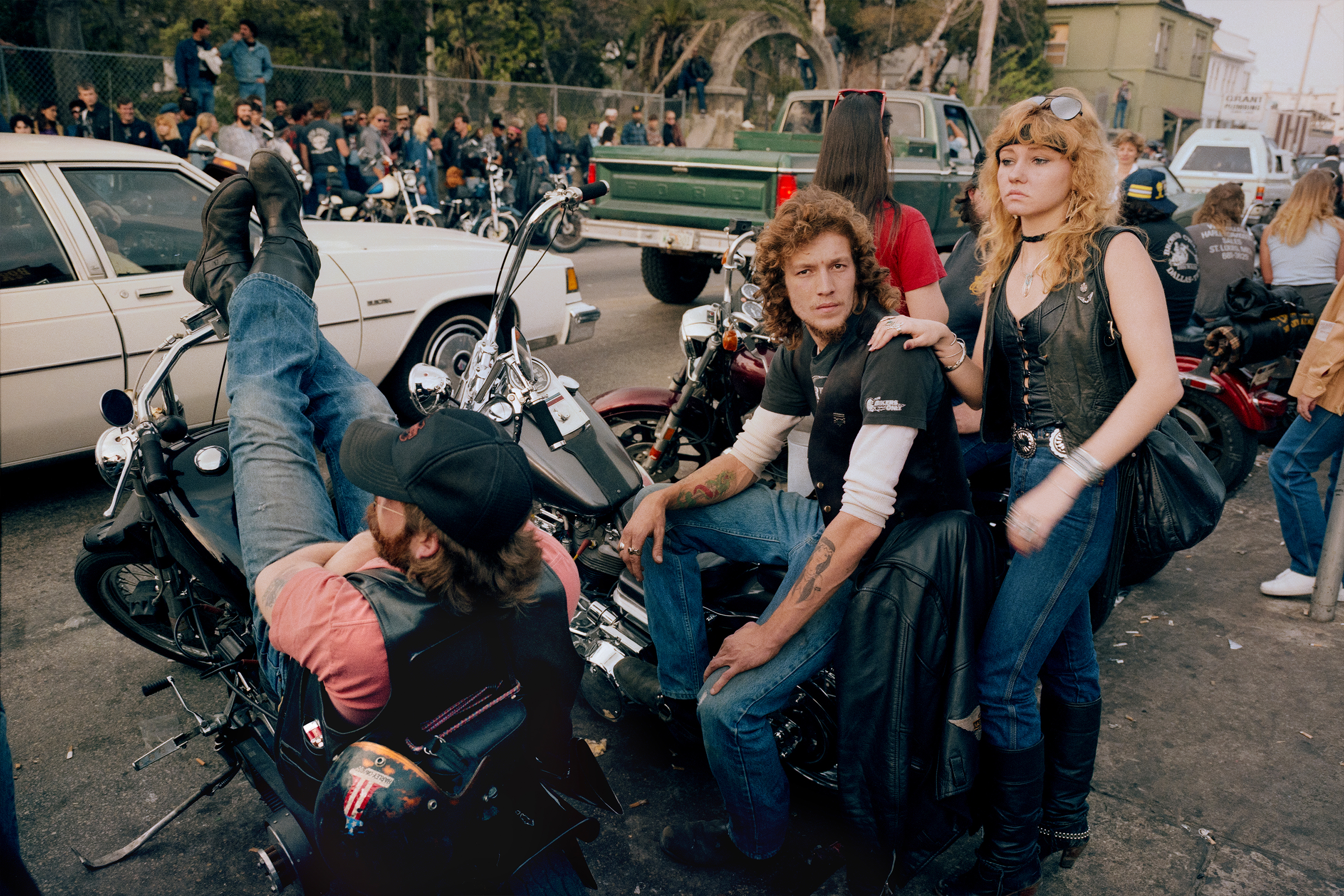Group of people in leather jackets and dark clothing gathered around motorcycles in street scene with cars and crowd in background.
