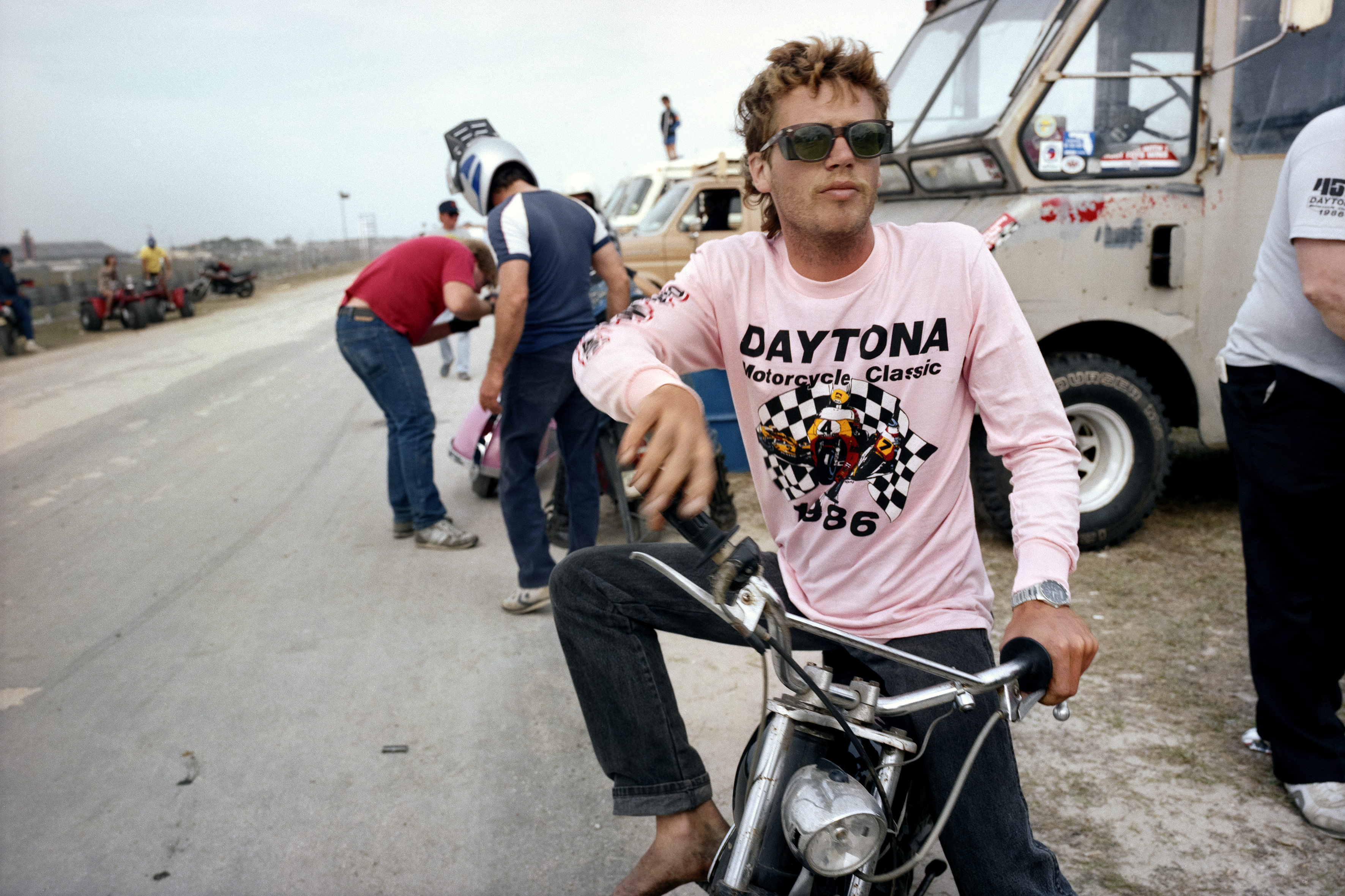 Man with curly hair and sunglasses wearing pink Daytona 1986 t-shirt sits on motorbike near white lorry with crew members working behind.