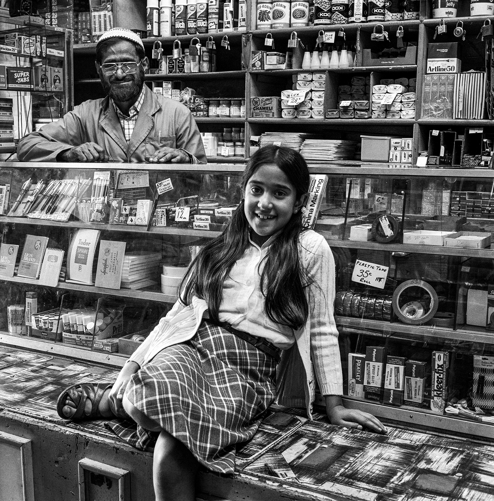 Black and white image of man with glasses behind bookshop counter and woman sitting cross-legged on floor amongst shelves of books.