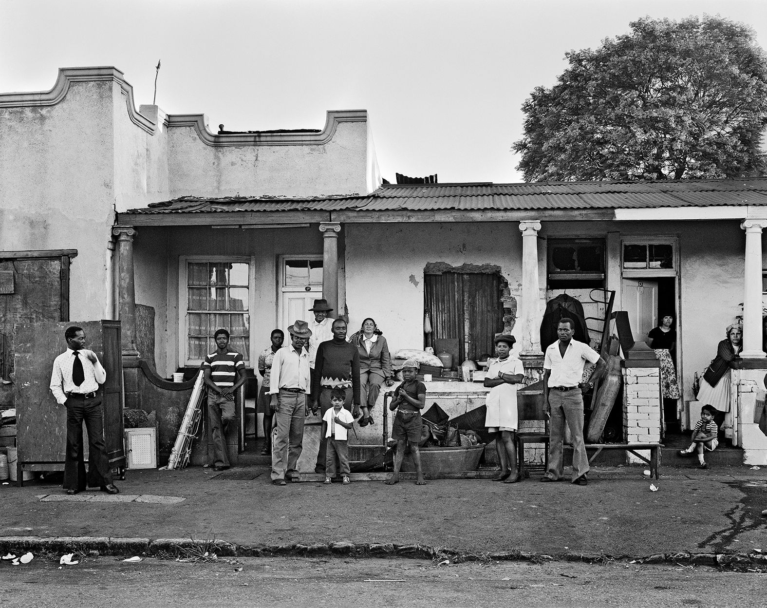 Black and white image showing people gathered outside a single-storey building with columns and large tree behind it.
