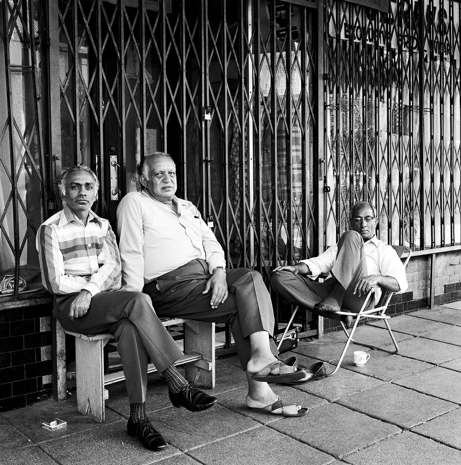 Three elderly men sitting on chairs outside metal security gates on tiled pavement. Black and white photograph.