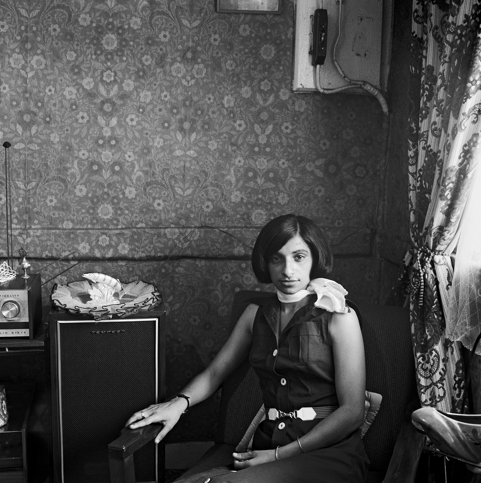 Black and white image of woman with bob haircut sitting in ornate Victorian parlour with patterned wallpaper and curtains.