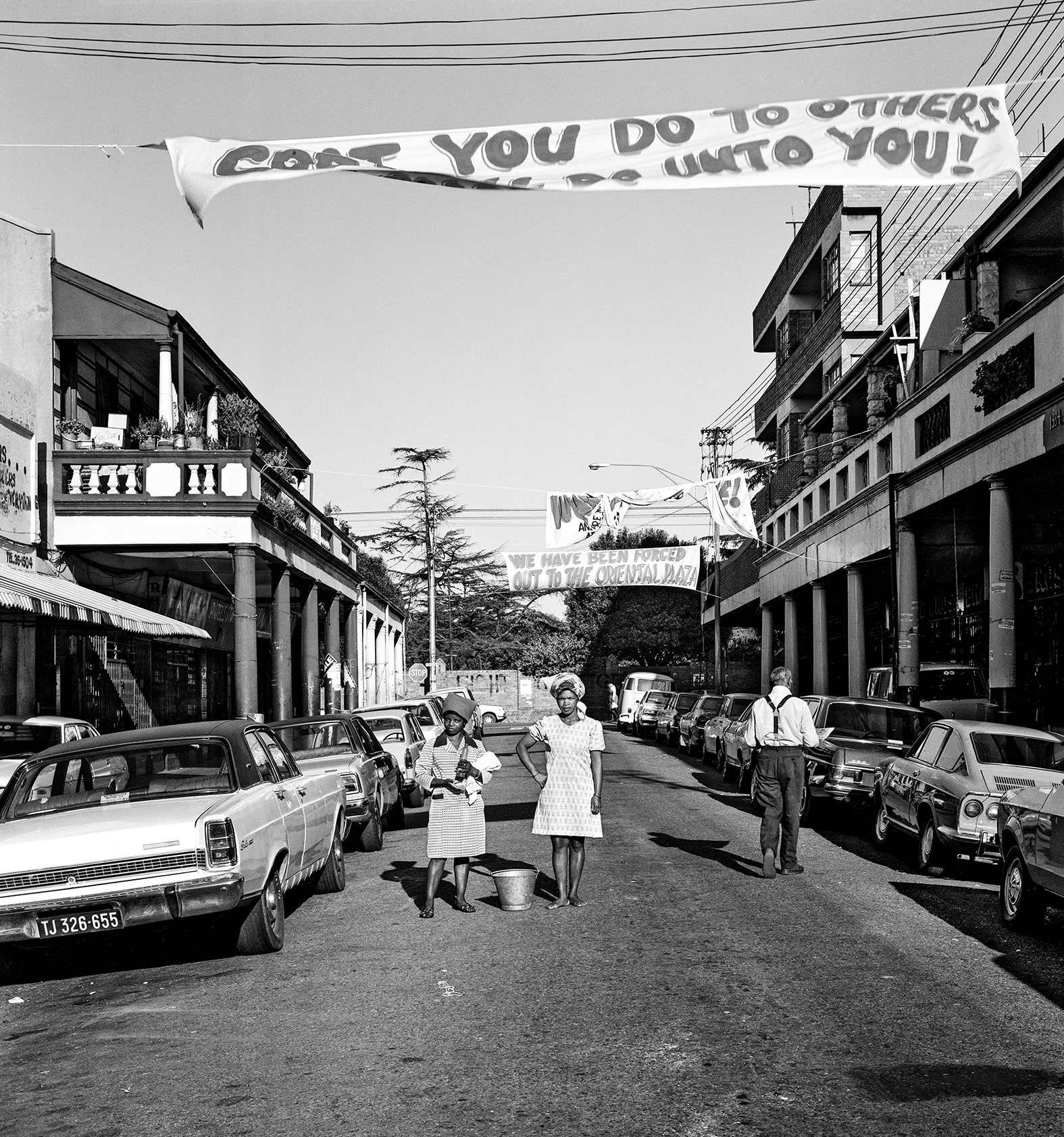 Black and white street scene with banner reading "YOU DO TO OTHERS AS THEY DO UNTO YOU!" spanning across road between buildings with vintage cars parked along sides.