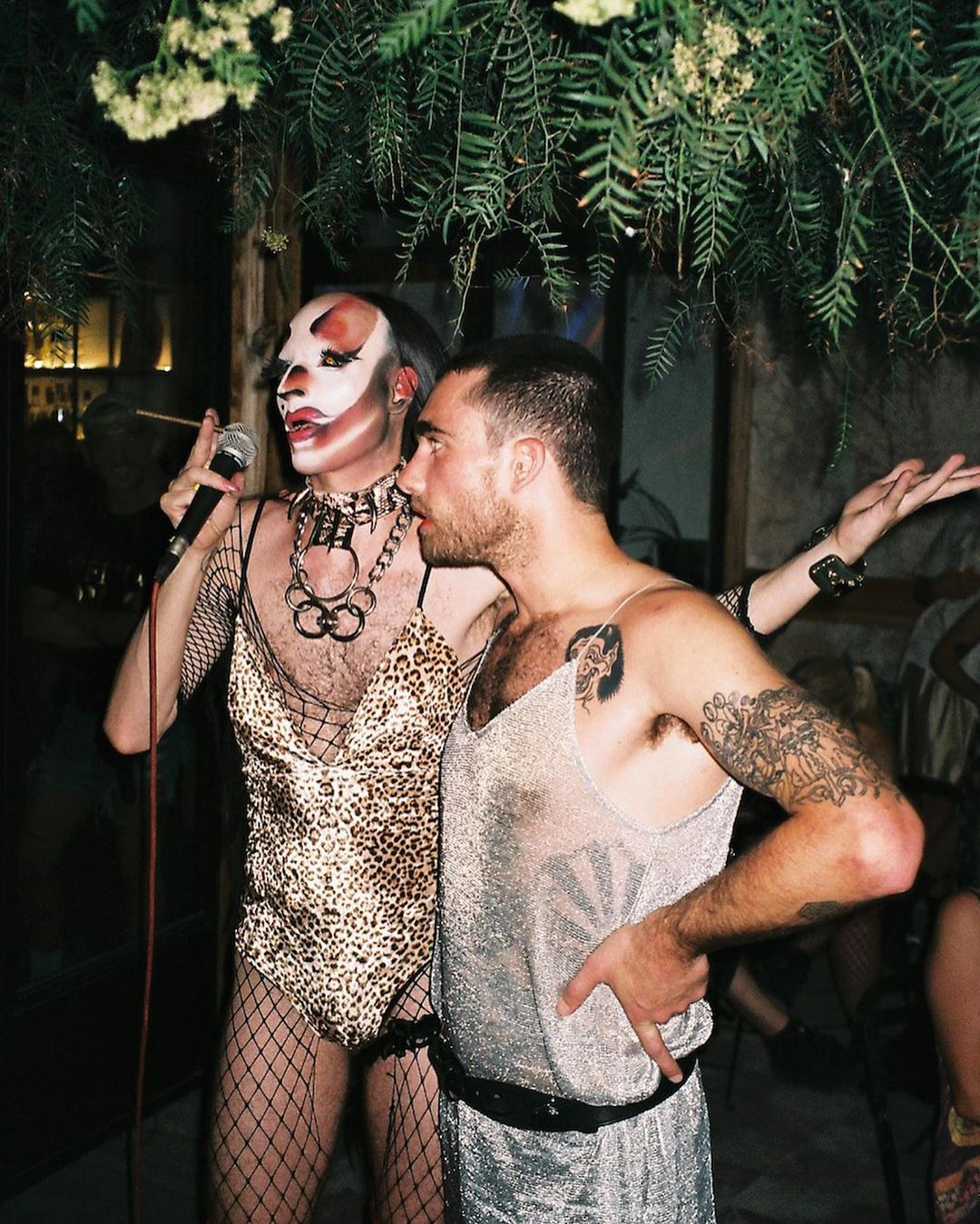 Drag performer in clown makeup and leopard print outfit singing into microphone whilst man in grey vest embraces them under greenery.