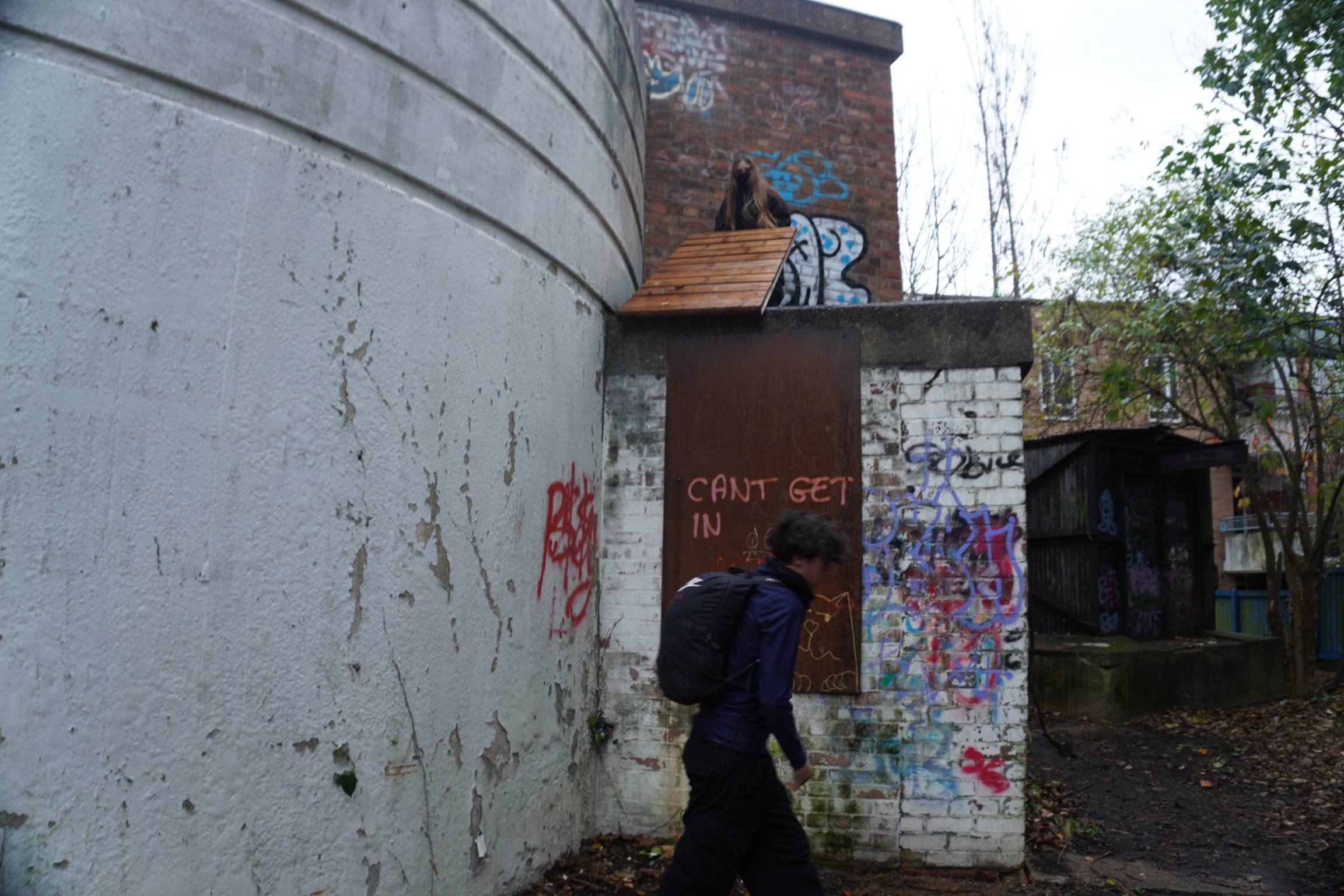 Person in dark clothing walking past abandoned industrial buildings with graffiti, surrounded by bare trees and overgrown vegetation.