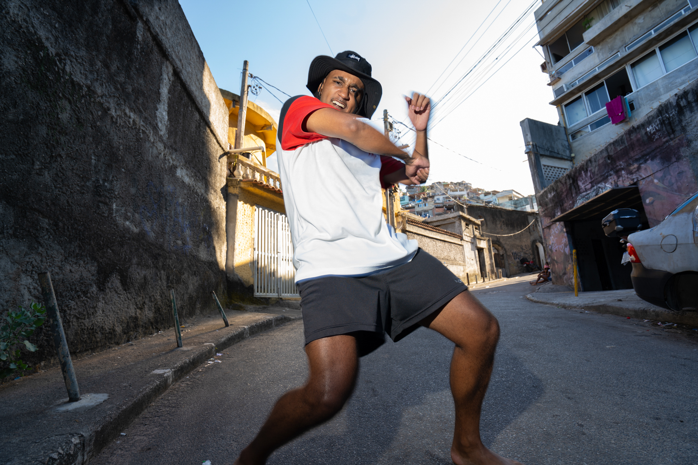 Man in white t-shirt and dark shorts dancing on concrete street between buildings, arms positioned as if holding something.