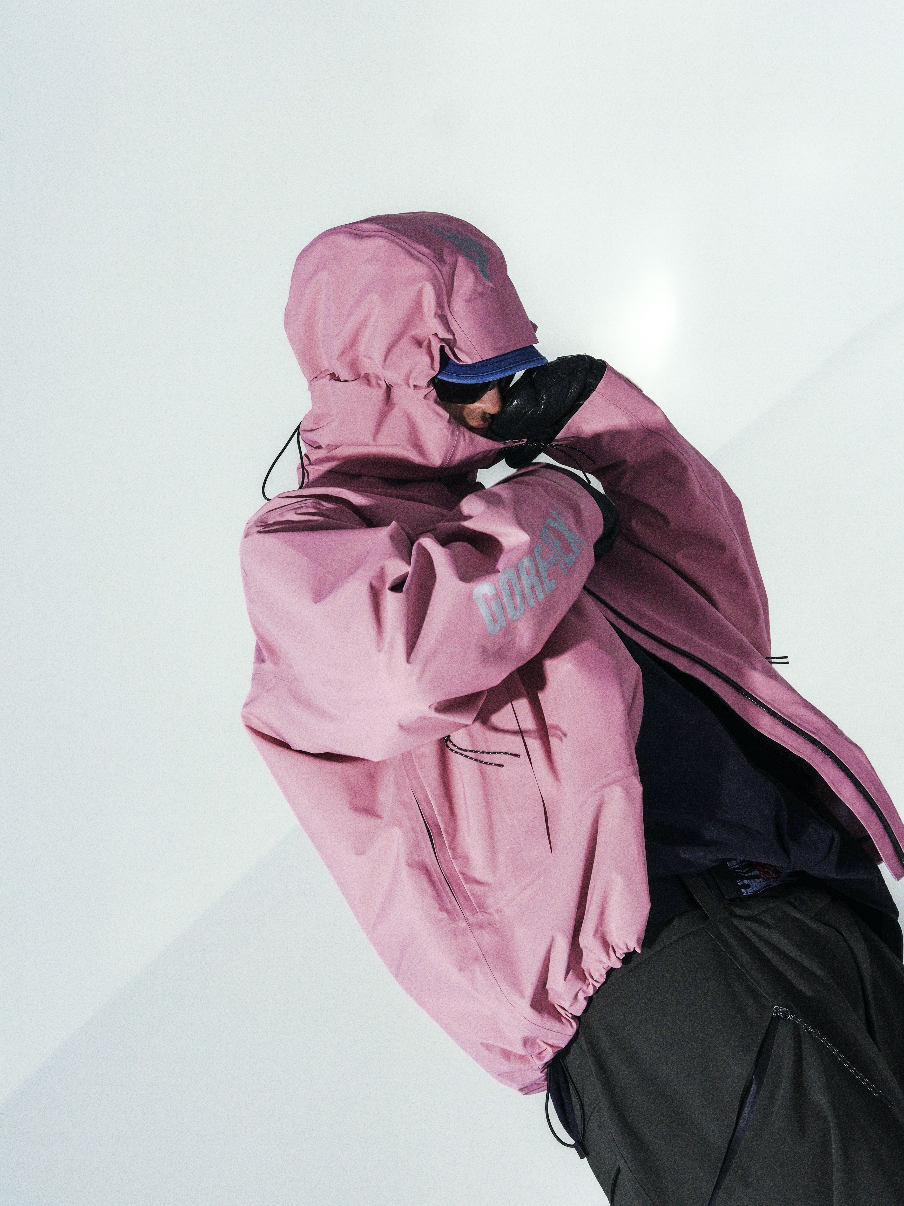 Person in pink hooded jacket and dark green trousers against white snowy background, face obscured by hood and dark sunglasses.