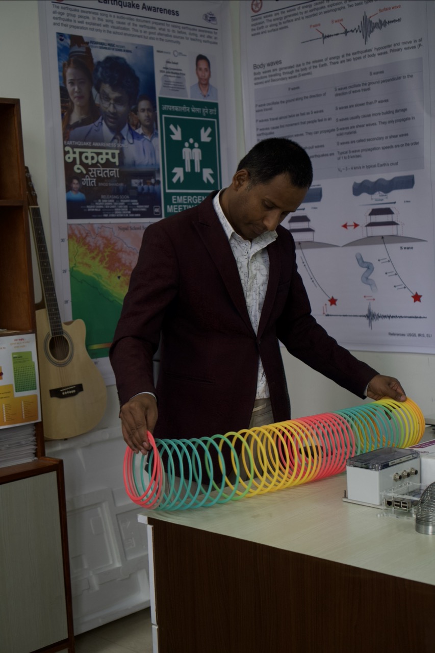 Man in dark blazer demonstrates colourful rainbow spring toy on white desk, with educational posters and charts on wall behind.