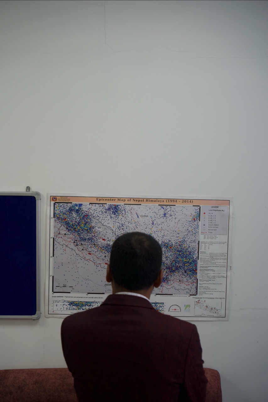 Person in dark suit viewed from behind studying a large wall-mounted map with coloured markings and text panels.